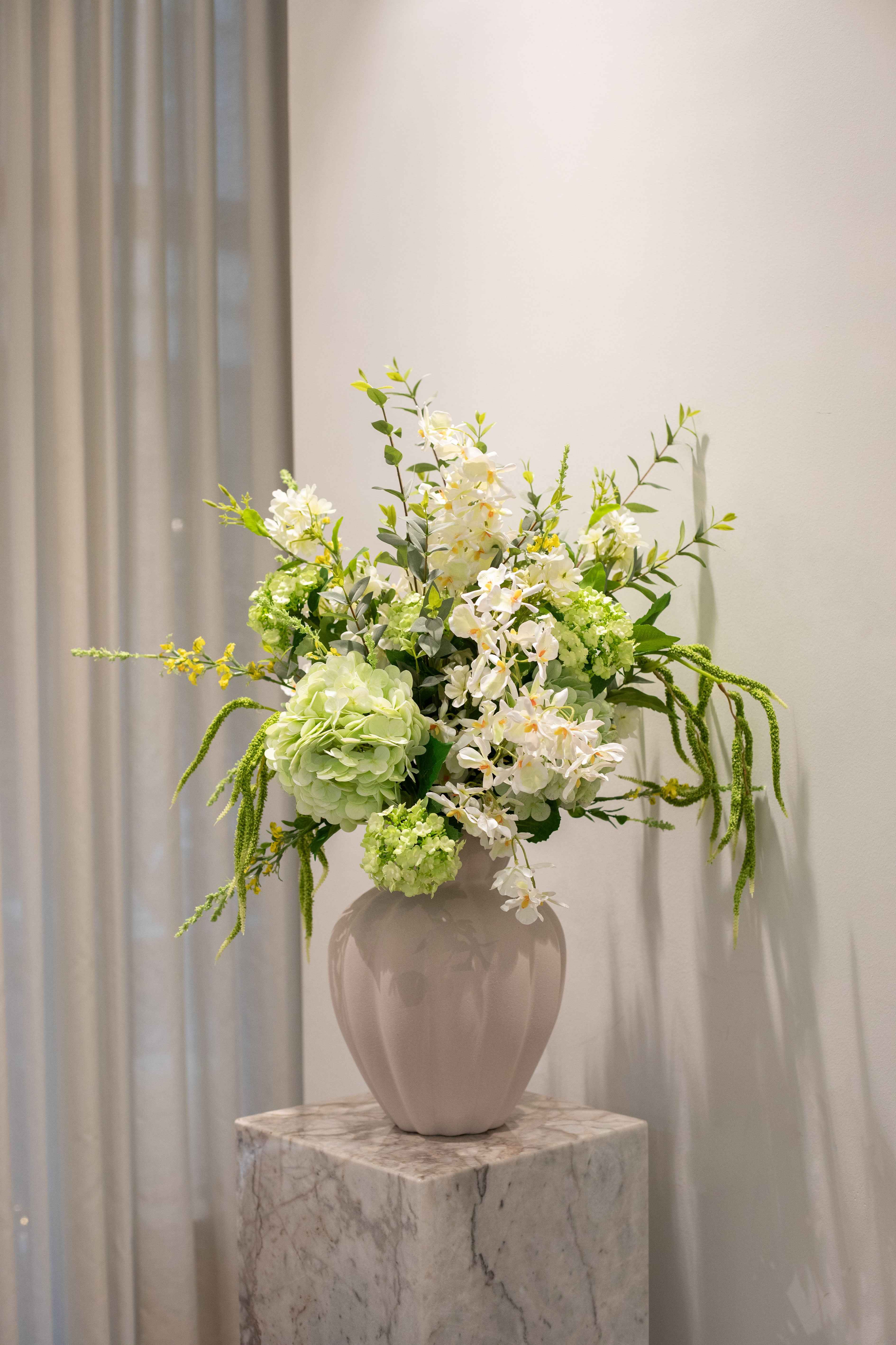 Kunstig blomster buket i hvide og grønne farver, der er placeret i en sand farvet keramik vase fra Specktrum. Vasen står på en marmor piedestal.