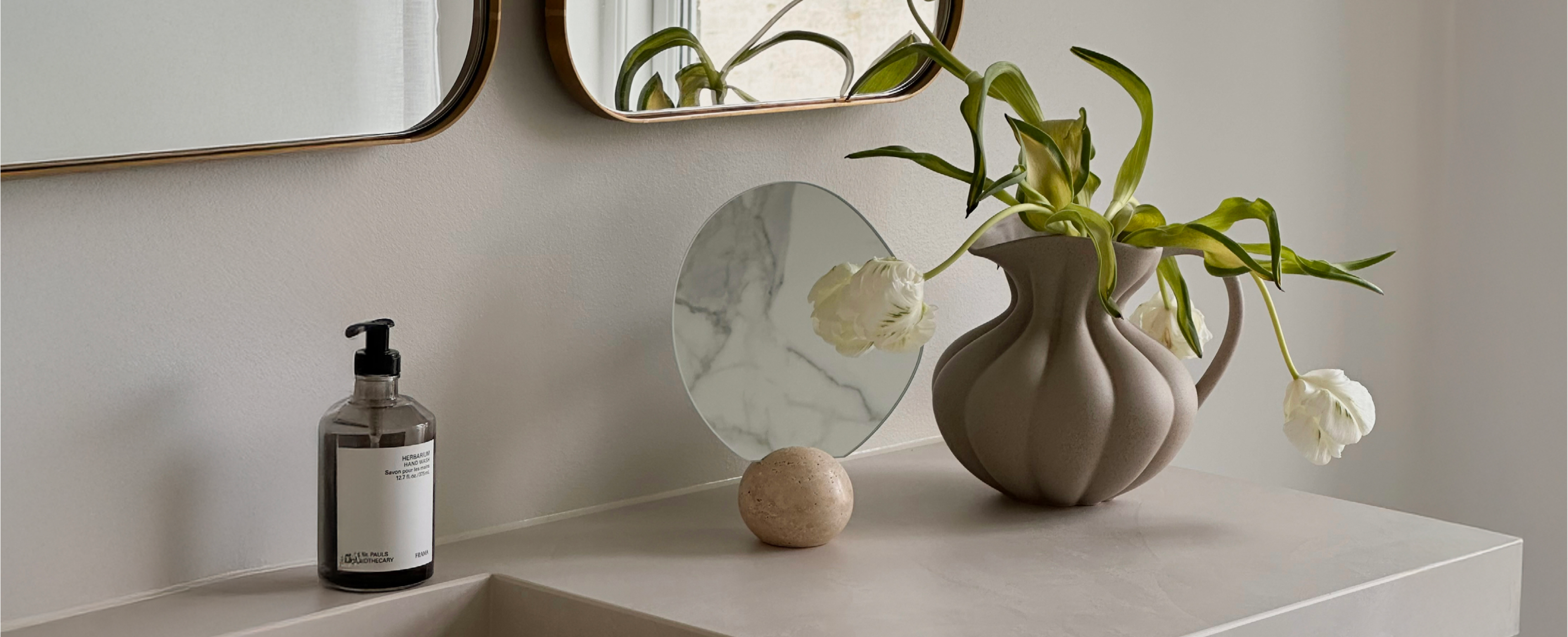 Asymmetrisk bordspejl med marmorfod, der står på et badeværelsesmøbel, ved siden af står en brun keramik kande med kunstige blomster.