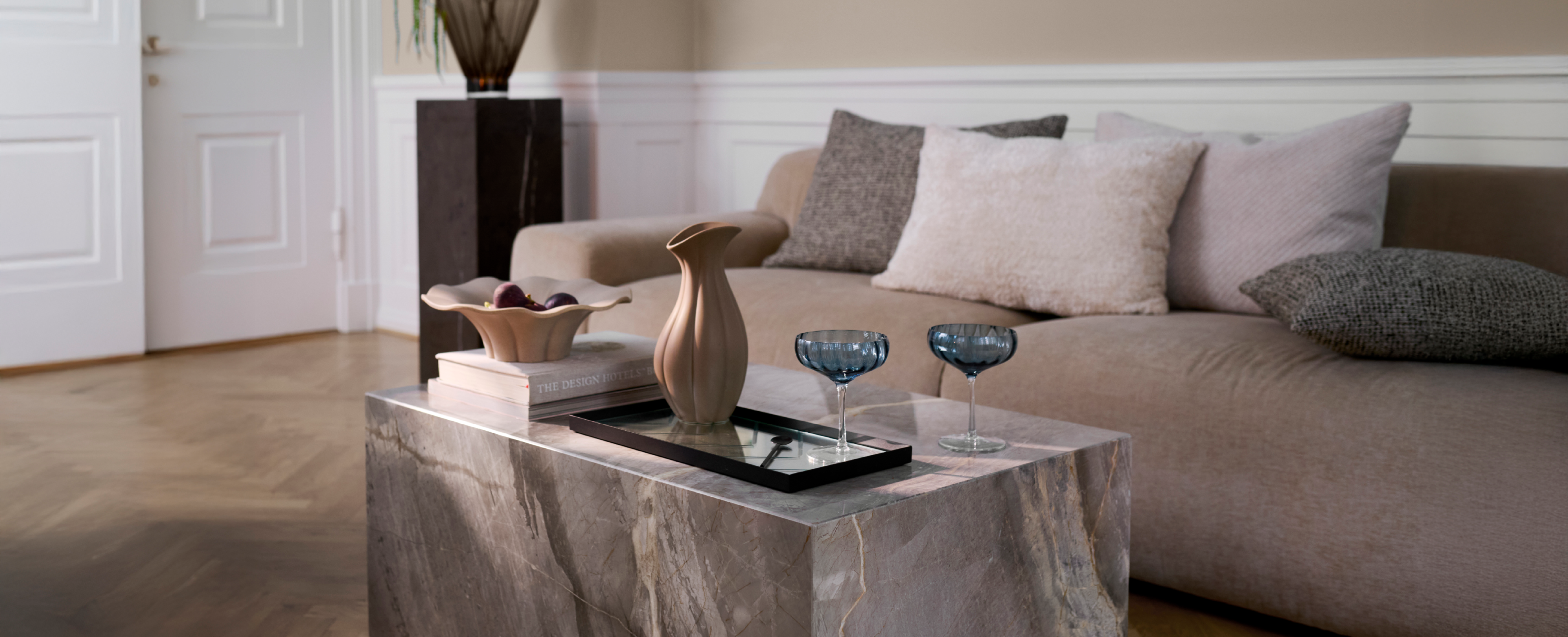 Marmor sofabord, hvorpå der står cocktail glas, en bakke med en keramik kande og et keramik fad ved siden af. Bag sofabordet er en brun sofa med puder i neutrale farver.