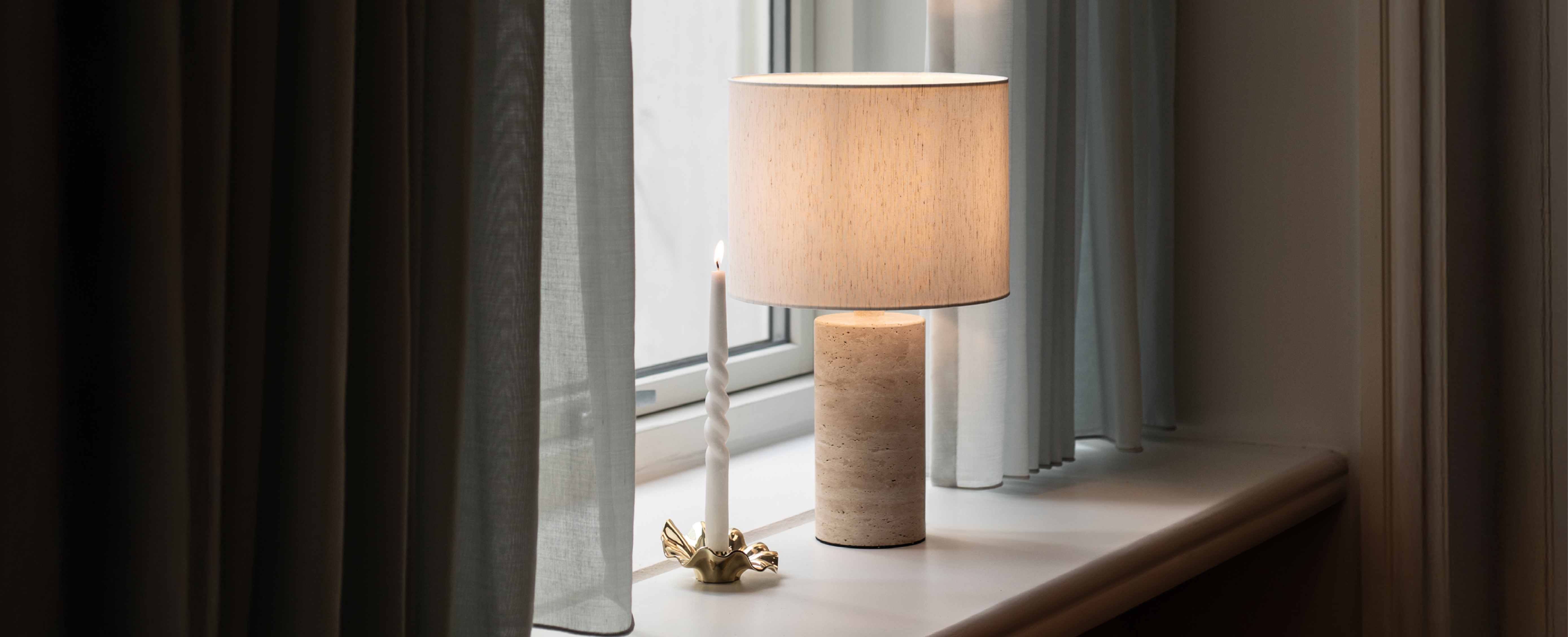 Tændt bordlampe med hvid lampeskærme fra Specktrum, der står i en vindueskarm. Ved siden af står en guld lysestage med hvidt snoede lys. Ved siden af vinduet hænger brune gardiner.