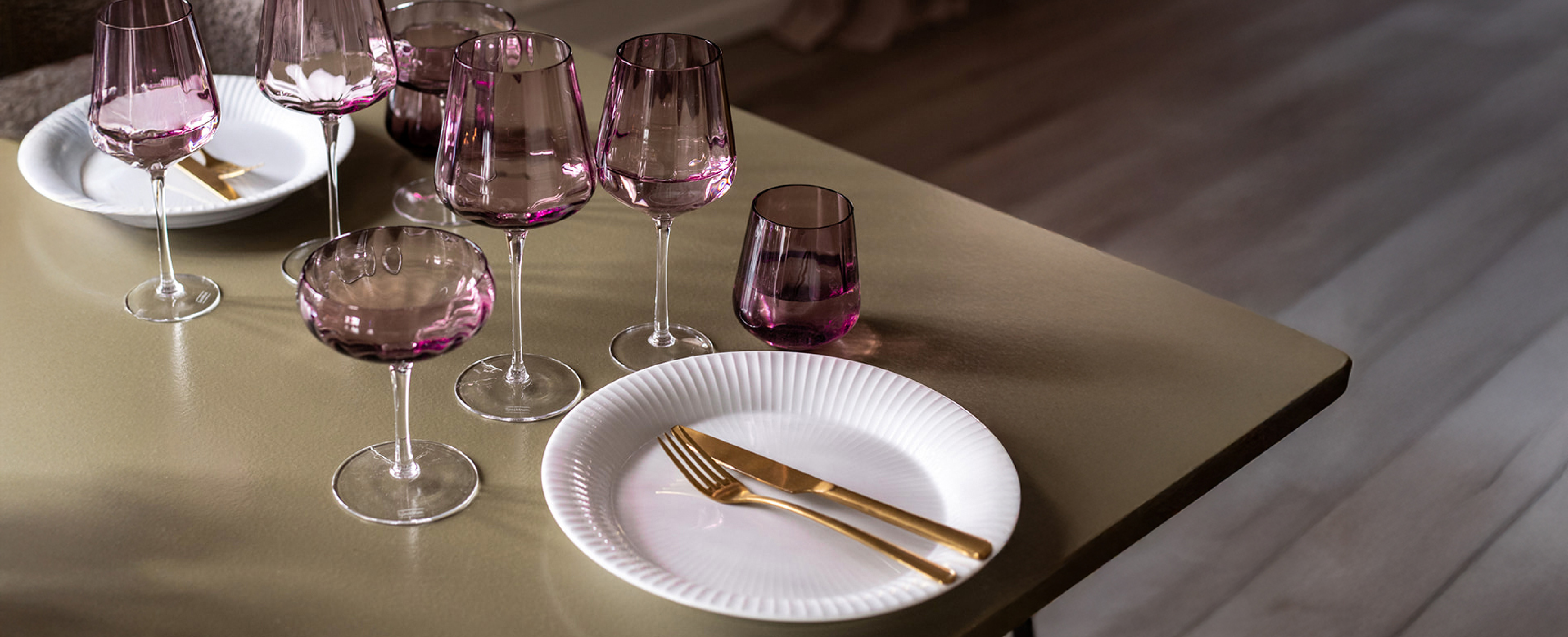 Grønt spisebord med rosa drikkeglas, vinglas og cocktail glas, samt hvide tallerkner og guld bestik.