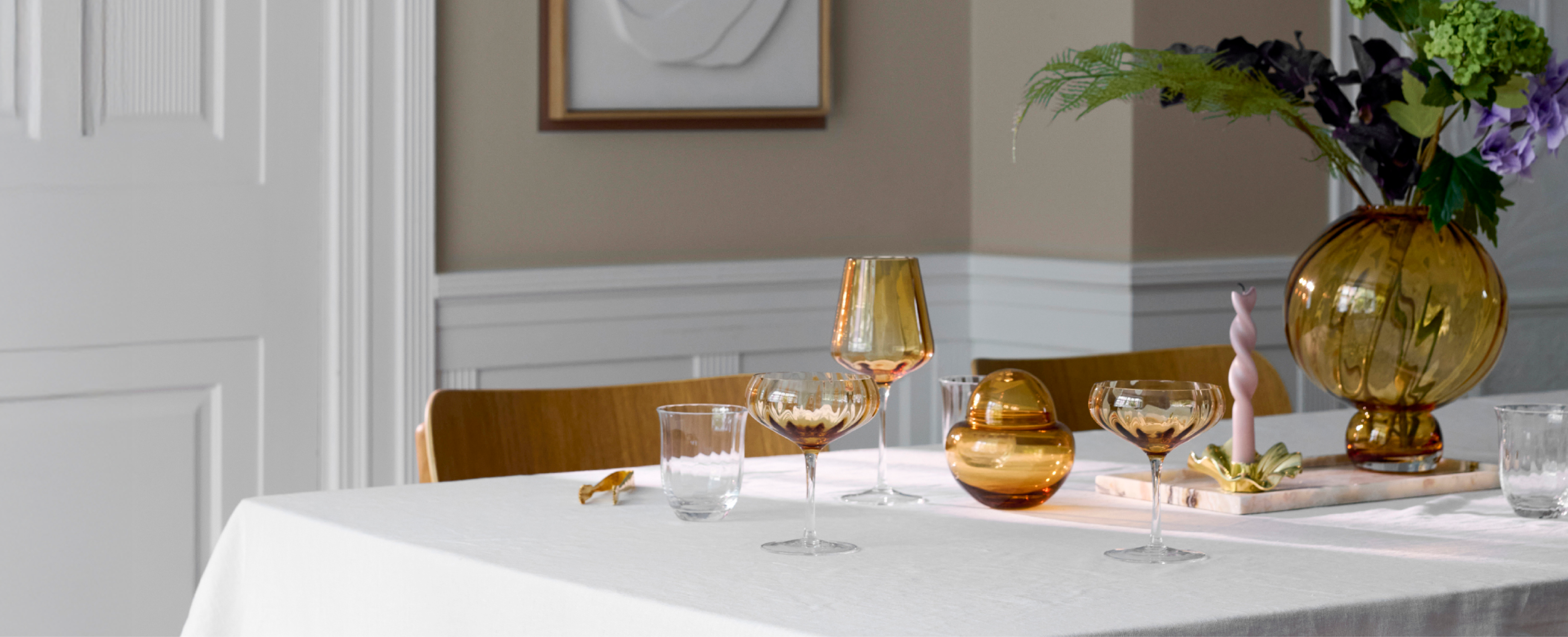 Billede af en borddækning med orange farvet glas, vaser og guld lysestage. Bordet er dækket med en hvid dug