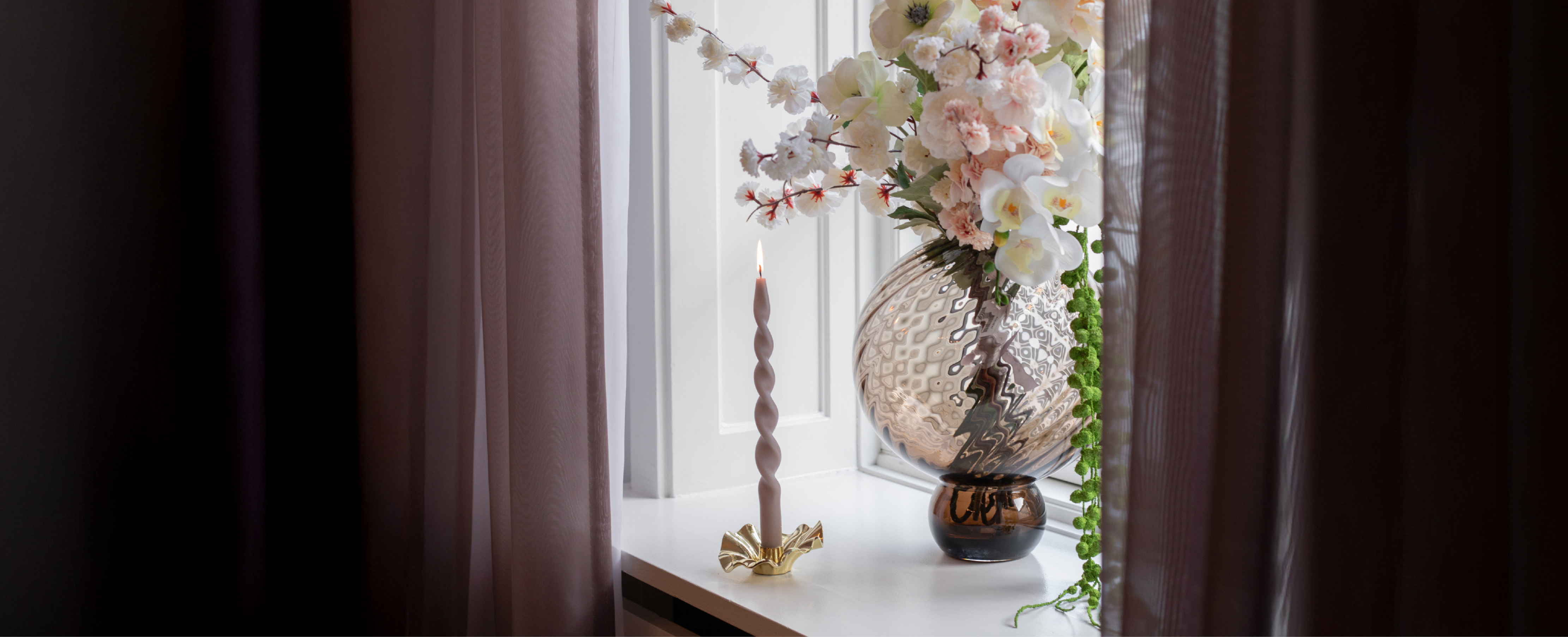 En vindueskarm hvor der står en stor brun rund glas vase med kunstige blomster i, ved siden af står en guld lysestage med et snoede lys. I siderne af vinduet hænger mørke gardiner.