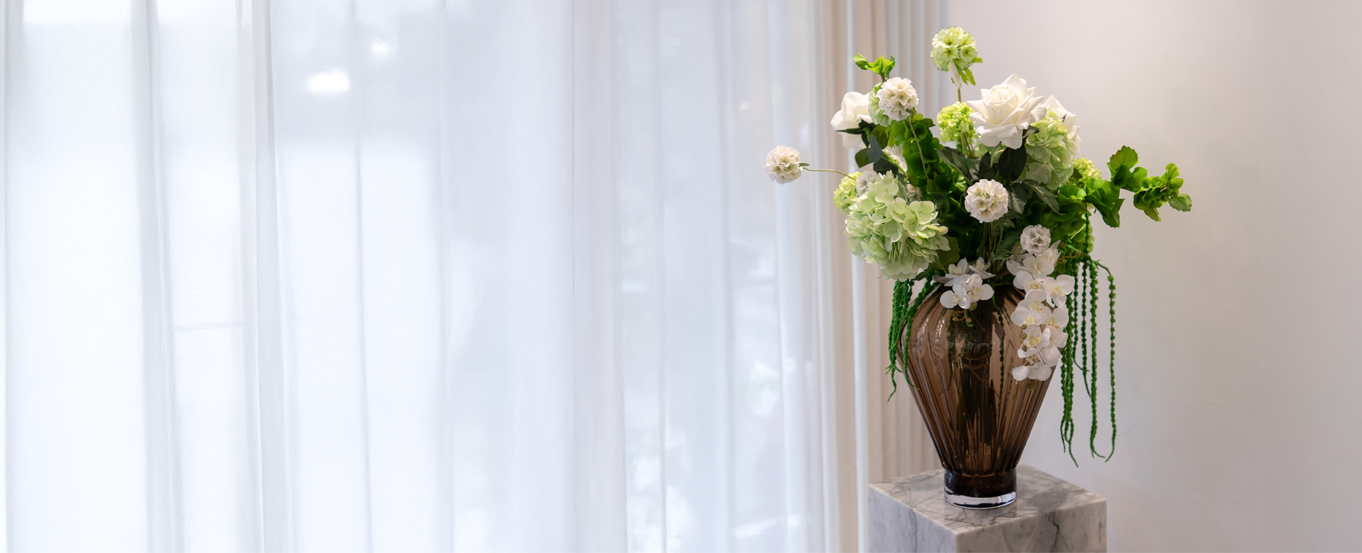 Buket af kunstige blomster i hvide og grønne farver, der er placeret i en røgfarvet glas vase. Blomsterarrangementet står på en marmor piedestal og billedet er taget foran en hvid væg og hvide gardiner.