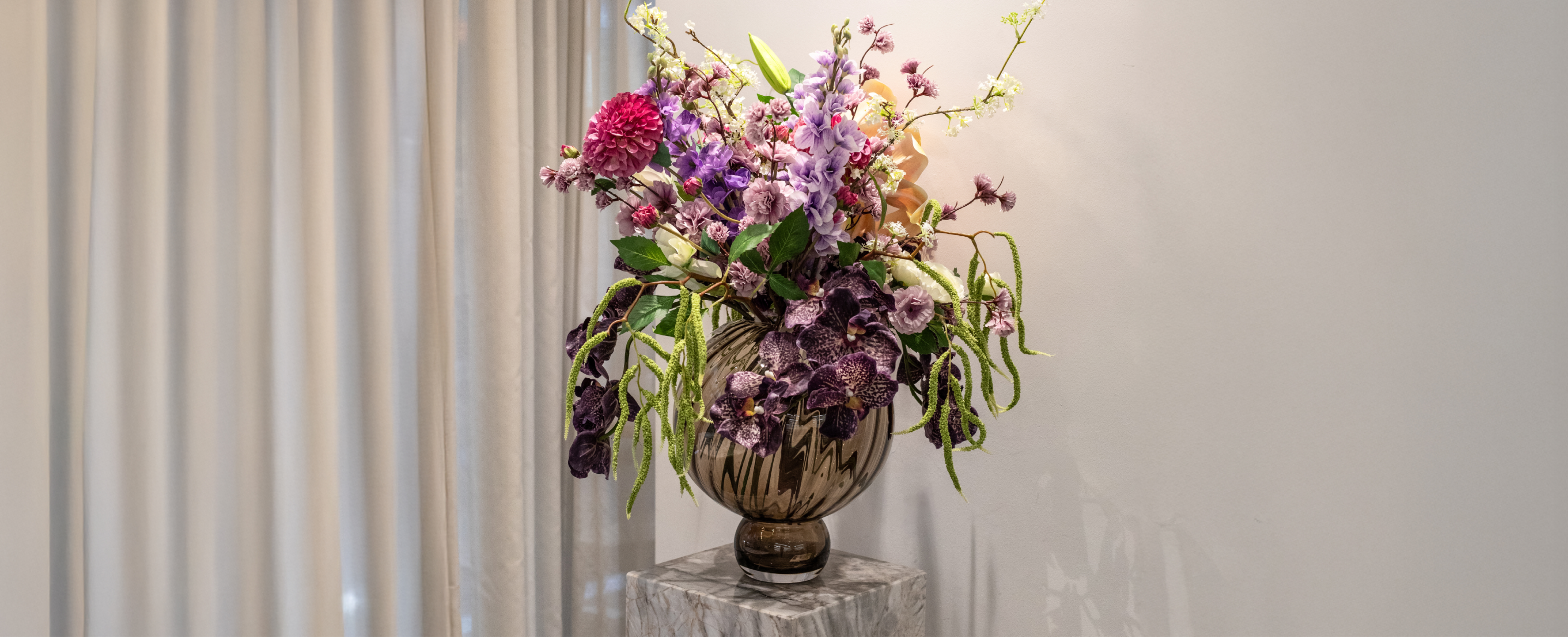 Stor kunstige buket i lilla, grønne og rosa nuancer, der står i røgfarvede glasvase på en marmor pedestal.