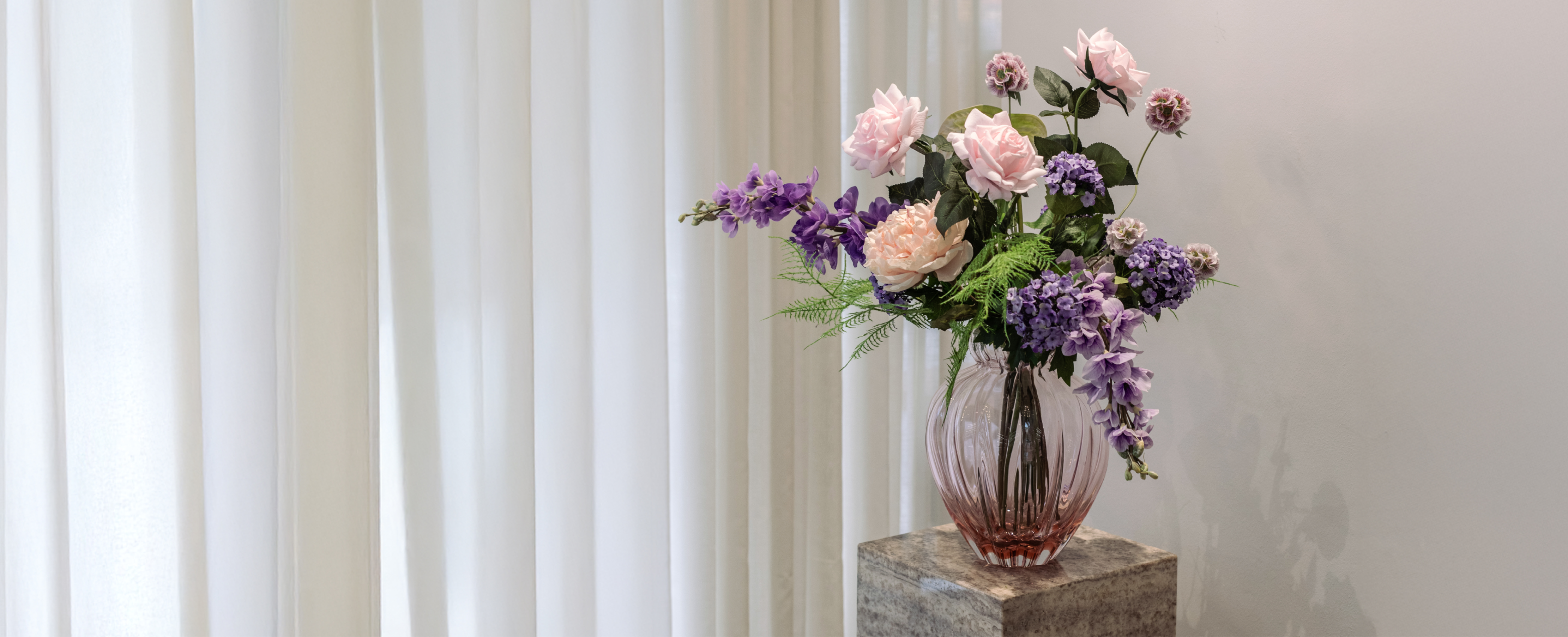 Kunstig buket i rose, lilla og grønne nuancer, der står i en lyserød glas vase oven på en marmor piedestal. Baggrunden er en hvid væg og hvide gardiner.