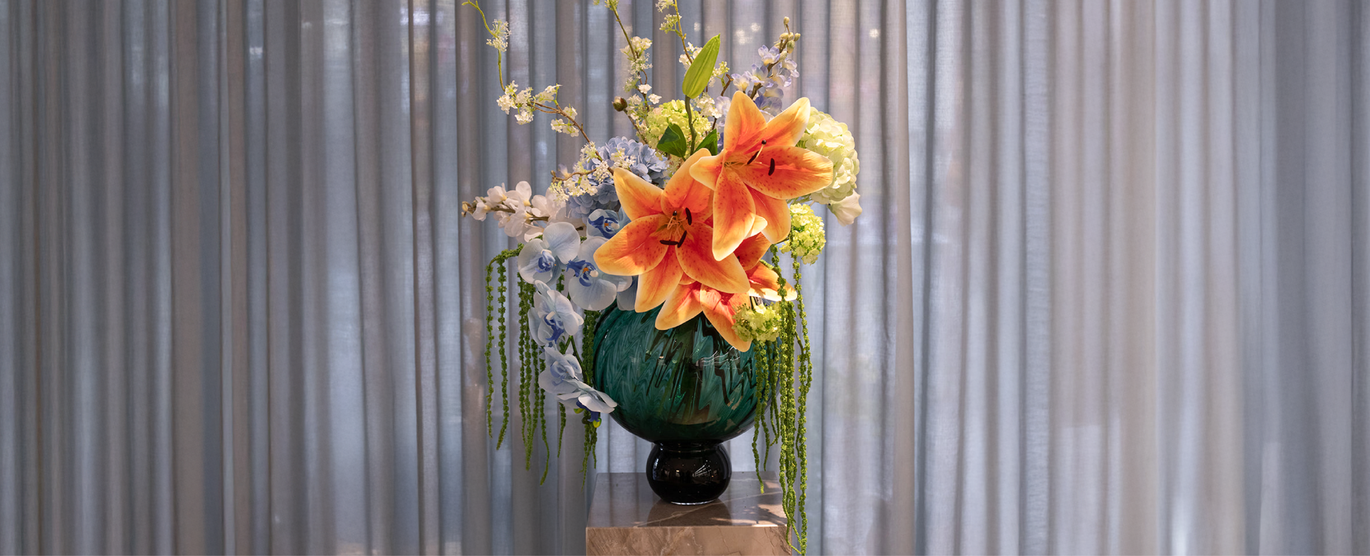 kunstig blomster buket i orange, grønne og blå nuancer, der er placeret i en mørkegrøn glas vase. Blomsterarrangementet står på en marmor piedestal, og baggrunden er blå gardiner.