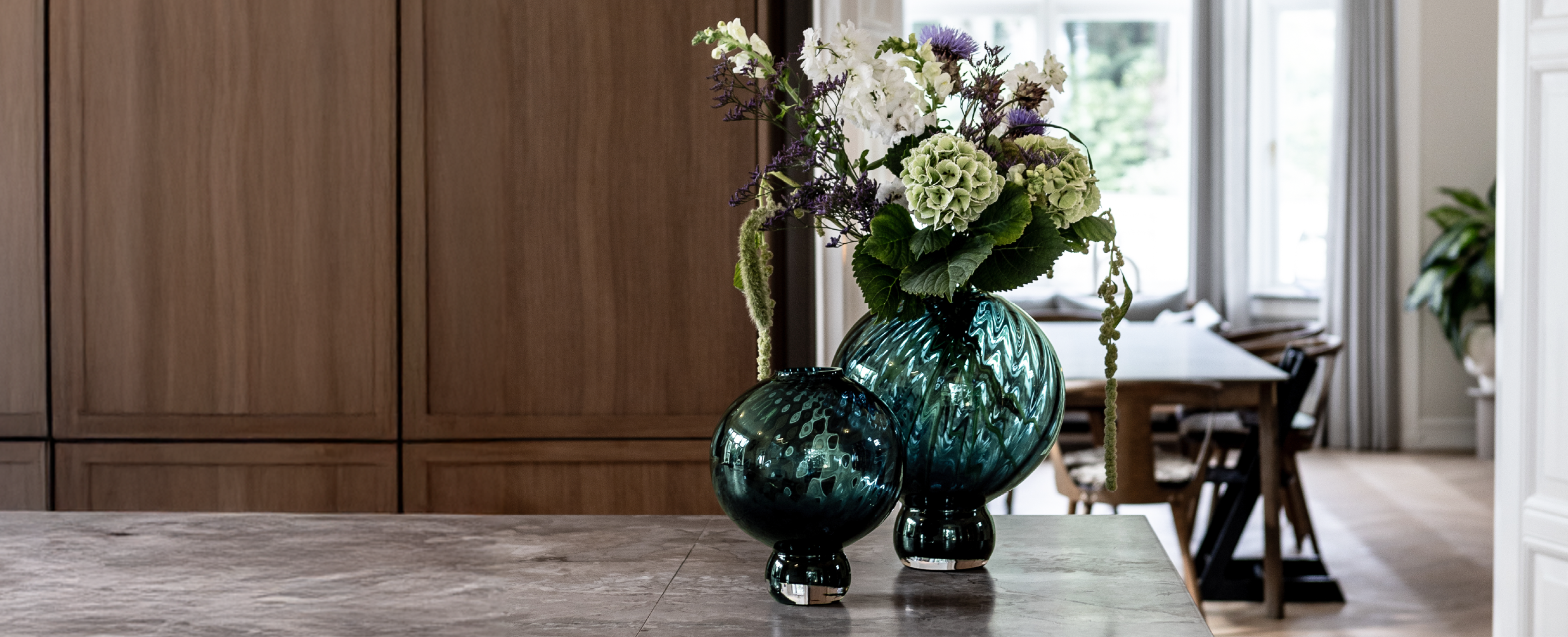 En stor og lille rund glasvase i grøn, der står på et køkkenbord. I den store vase er en buket kunstige blomster. Baggrunden er brune træ skabe og en spisestue, med et spisebord og stole.