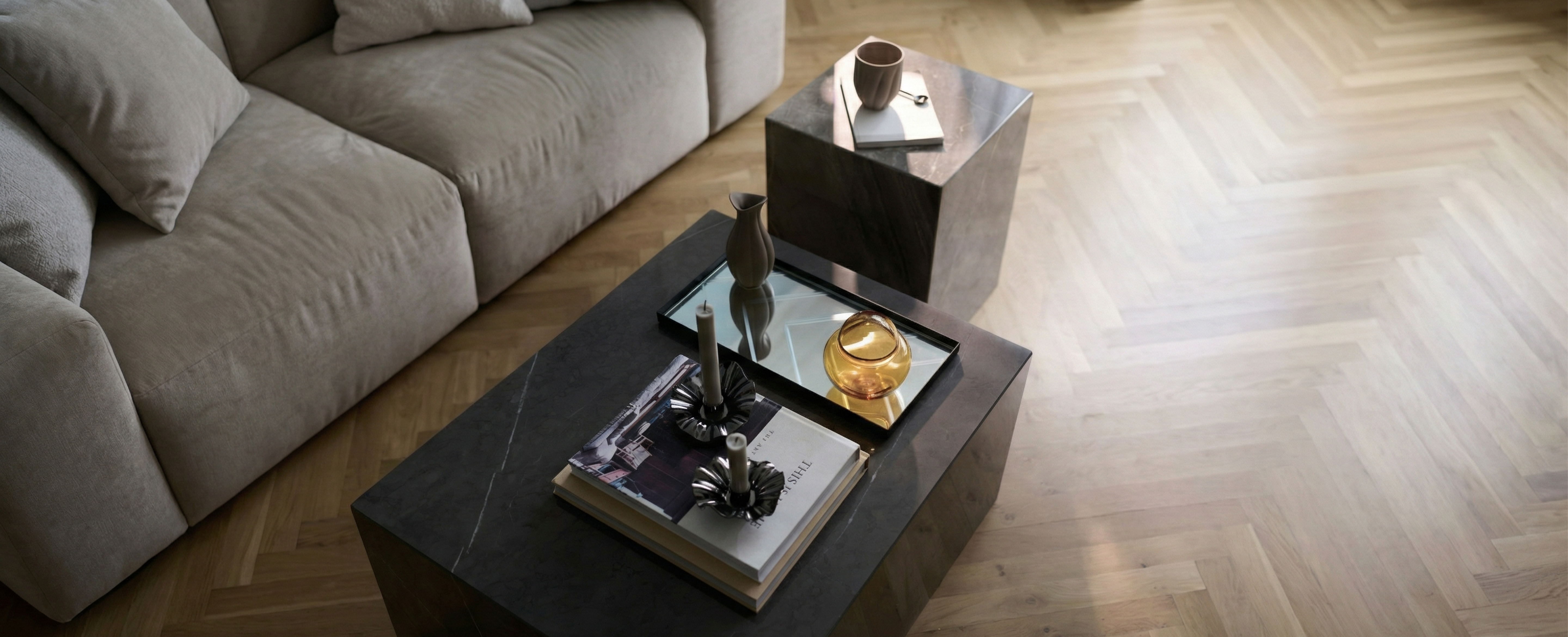 Marmor sofabord hvorpå der ligger magasiner, en spejlbakke med en kande og bonbonniere og metal lysestager med lys. Ved siden af står et matchende sidebord med en kaffekop. Bagved er en brunlig/beige sofa. Gulvet er træ sildeben.