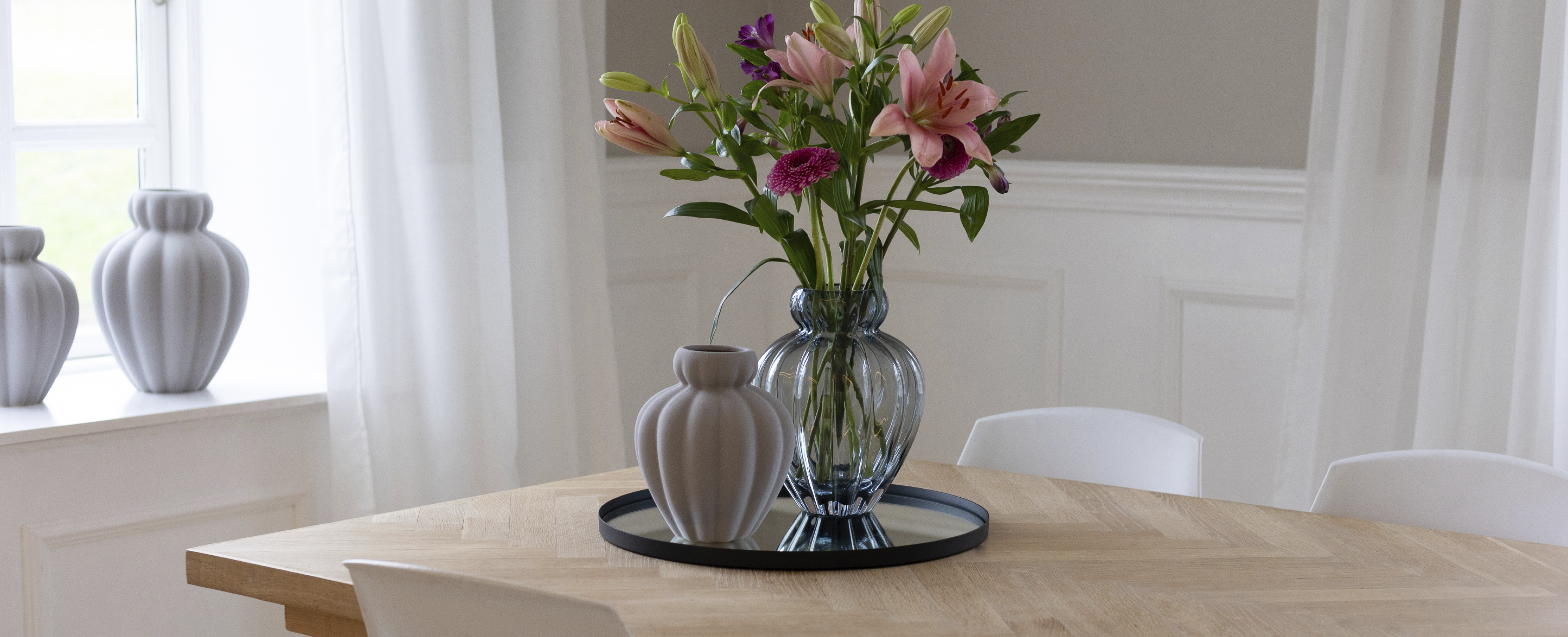 Spisebord i træ med en glasvase med lyserøde blomster på en sort bakke, i et lyst rum med hvide stole og blødt naturligt lys.