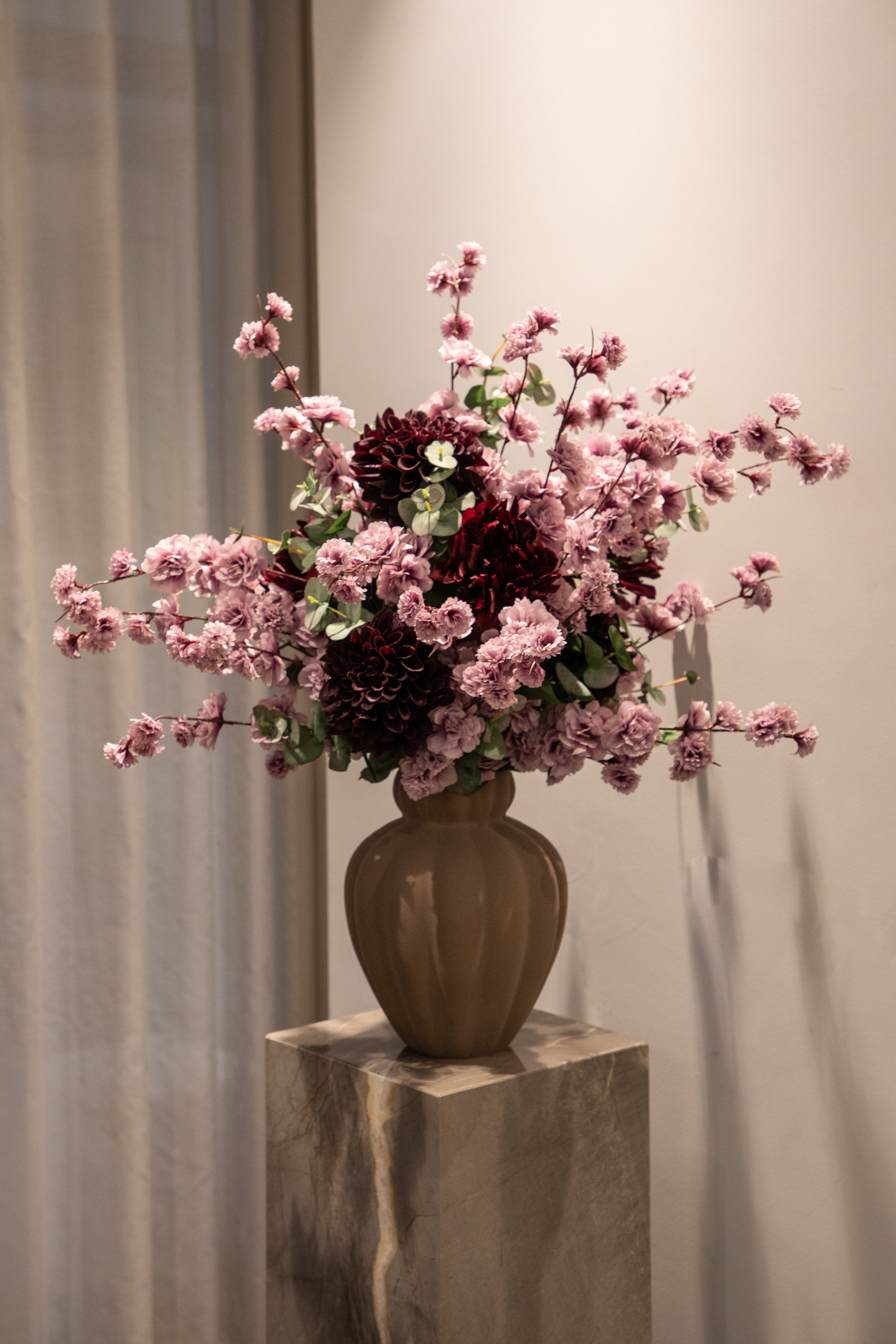 Stor kunstig buket i rose, grønne og bordeaux nuancer, der står i en brun keramik vase, placeret ovenpå en marmor piedestal.