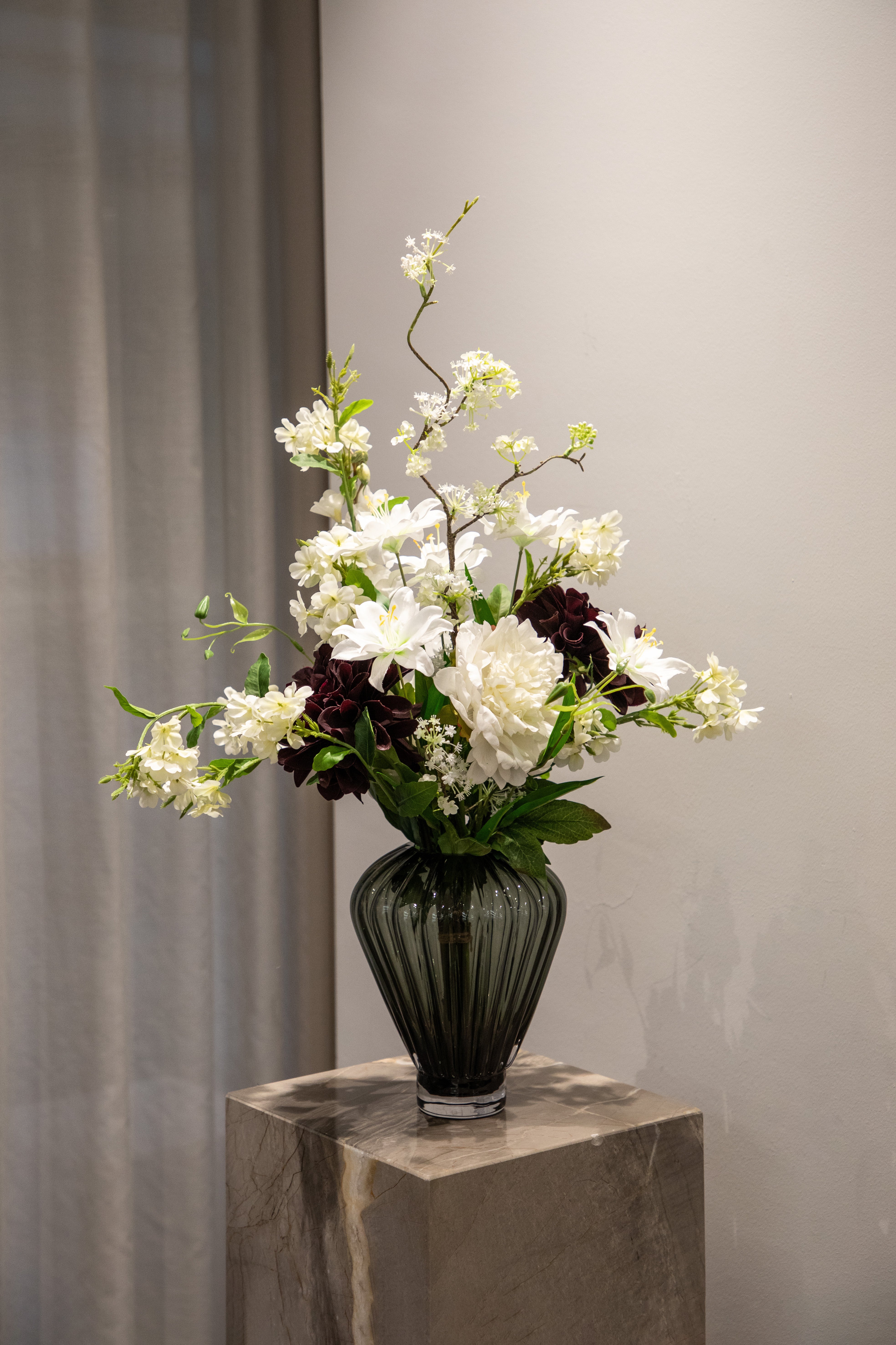 Kunstig buket i hvide, grønne og bordeaux nuancer, placeret i en høj grå glas vase, der står på en marmor piedestal.