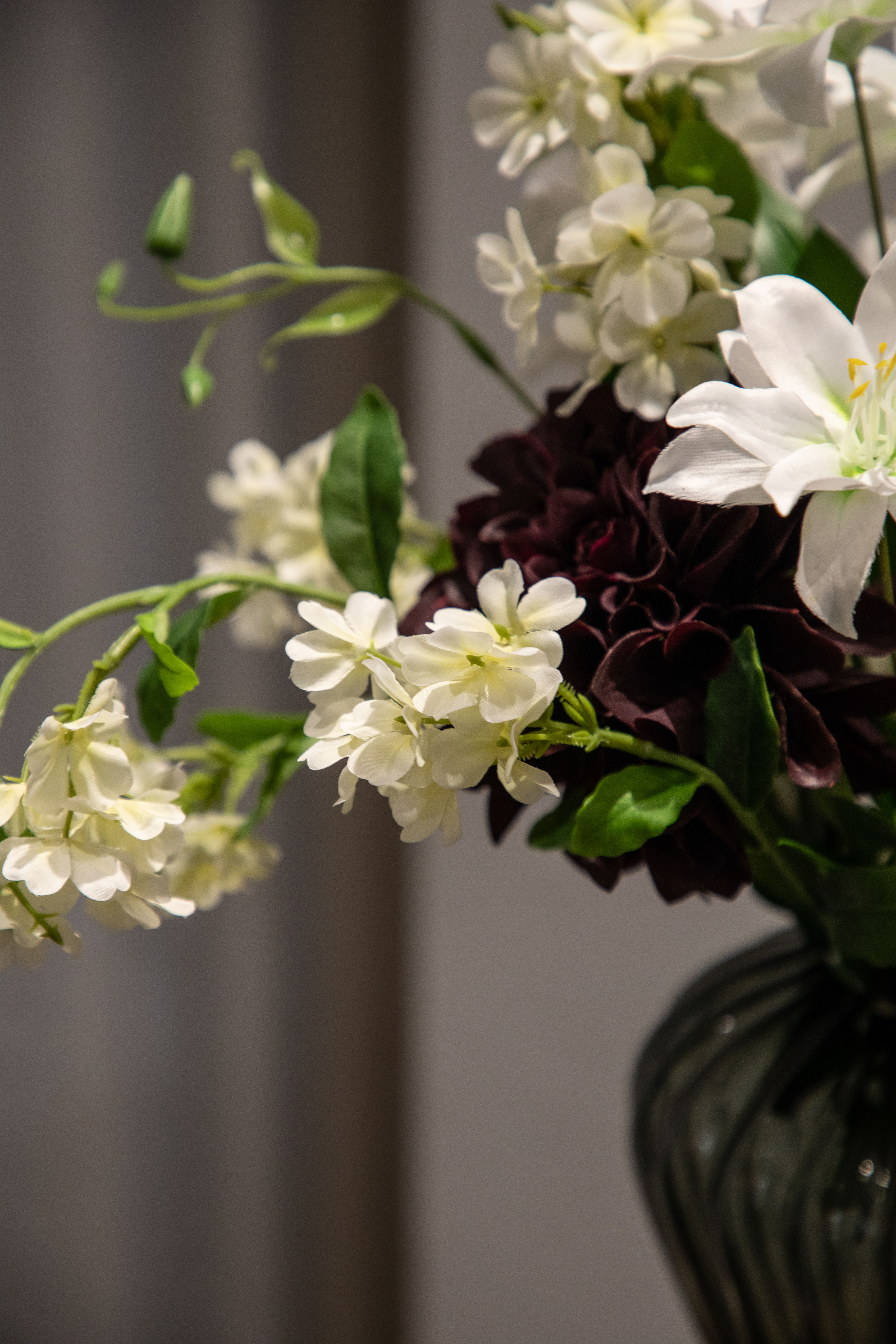 Nærbillede af hvide fontanesia stilke, der del af en større kunstig buket i hvide, grønne og bordeaux nuancer, buketten står i en grå glas vase.