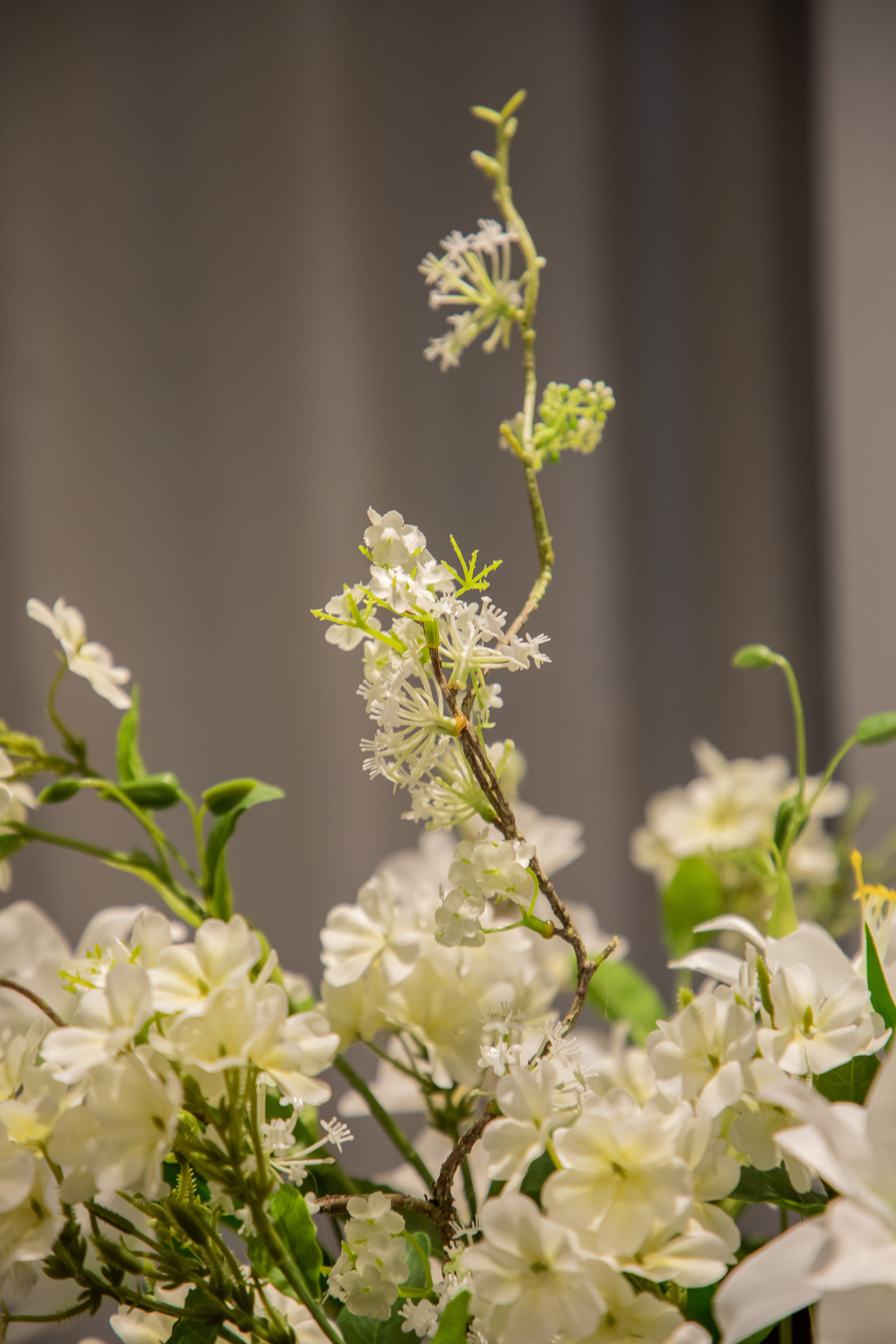 Nærbillede af hvid fontanesia stilk, der stikker op fra en større kunstig buket i hvide, grønne og bordeaux nuancer.