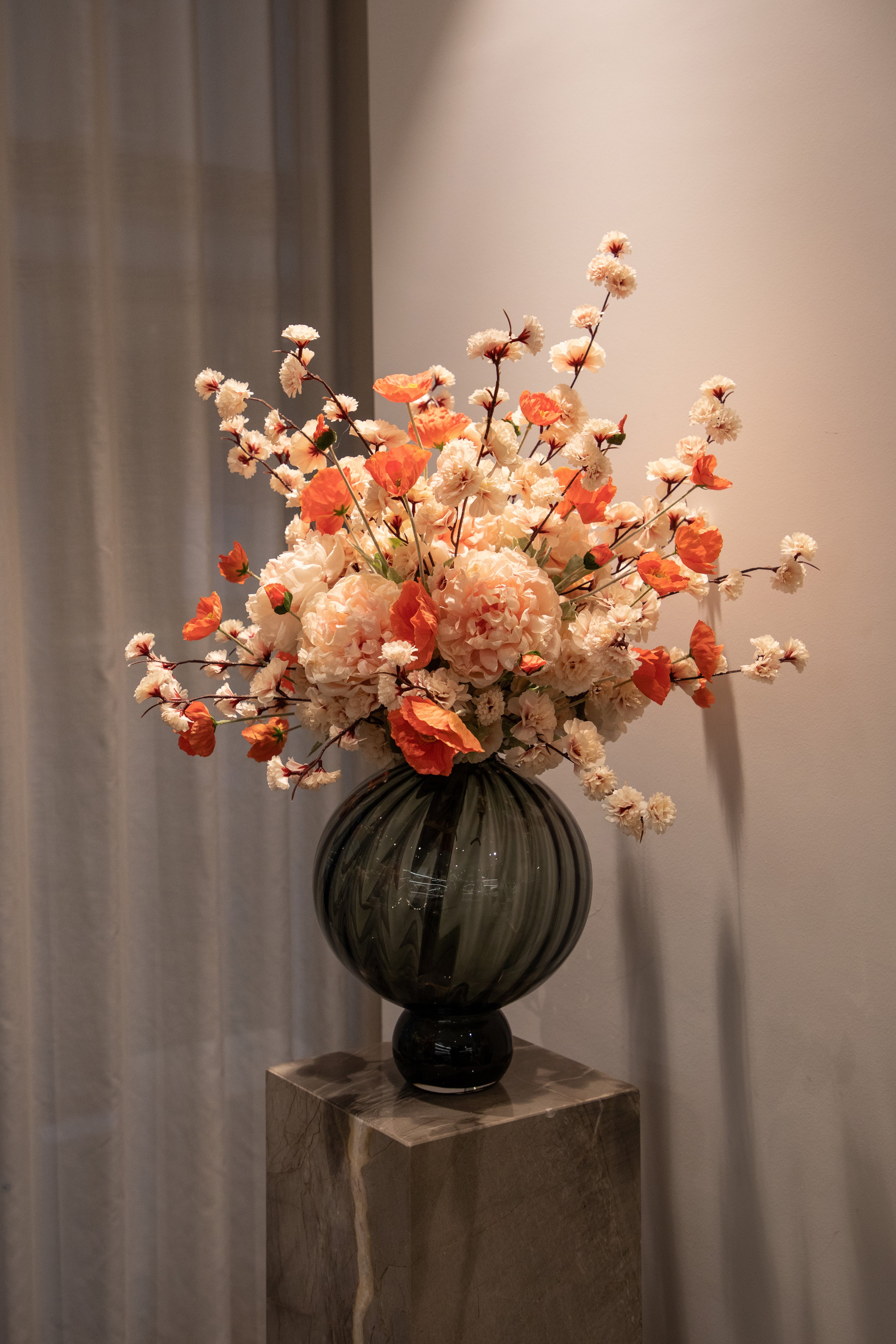 Stor kunstig buket i sarte rose og orange nuancer, der er placeret i en rund grå glas vase, der står ovenpå en marmor piedestal.