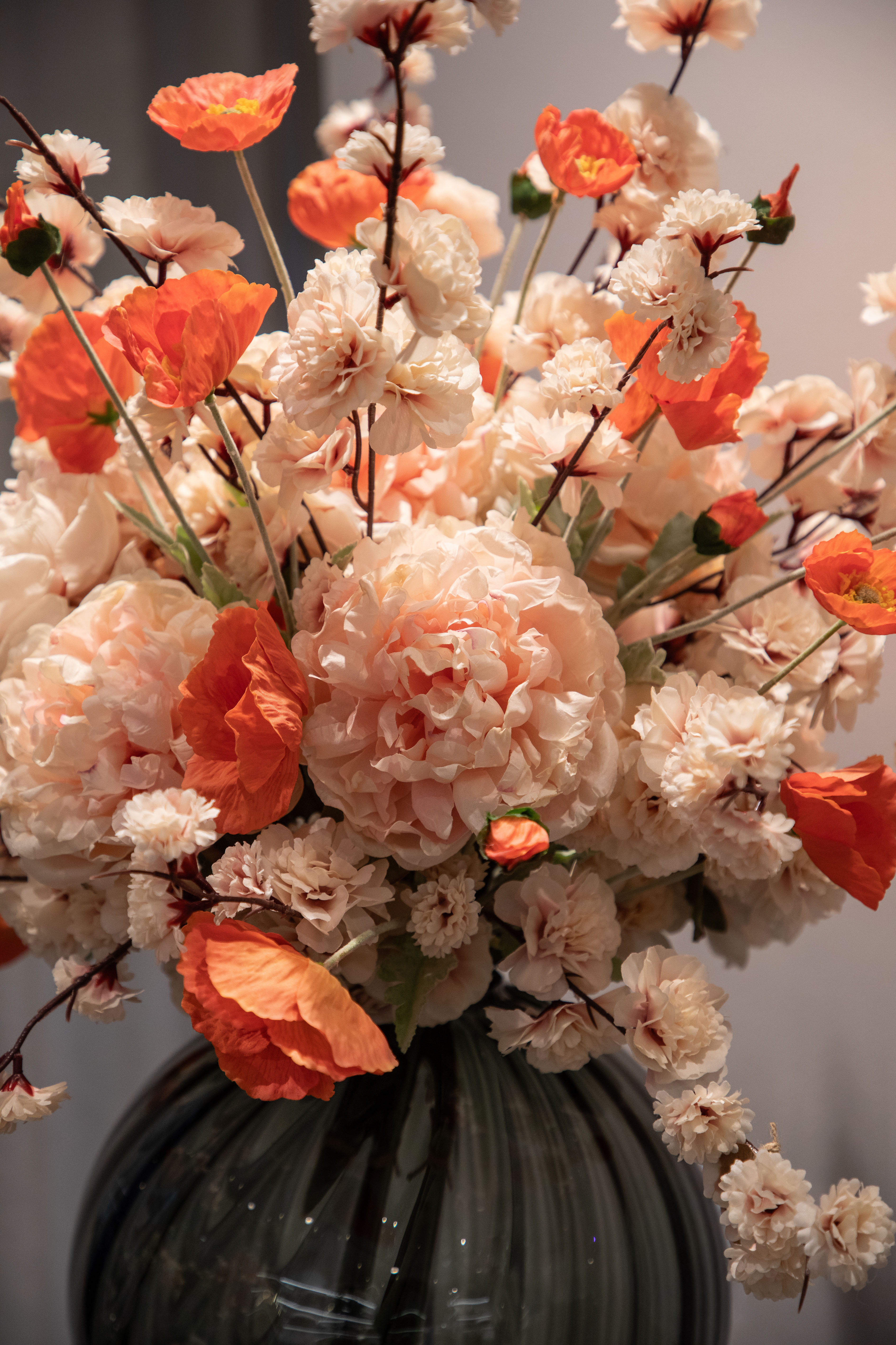 Nærbillede af stor kunstig buket i rose og orange nuancer, placeret i en rund grå glas vase.