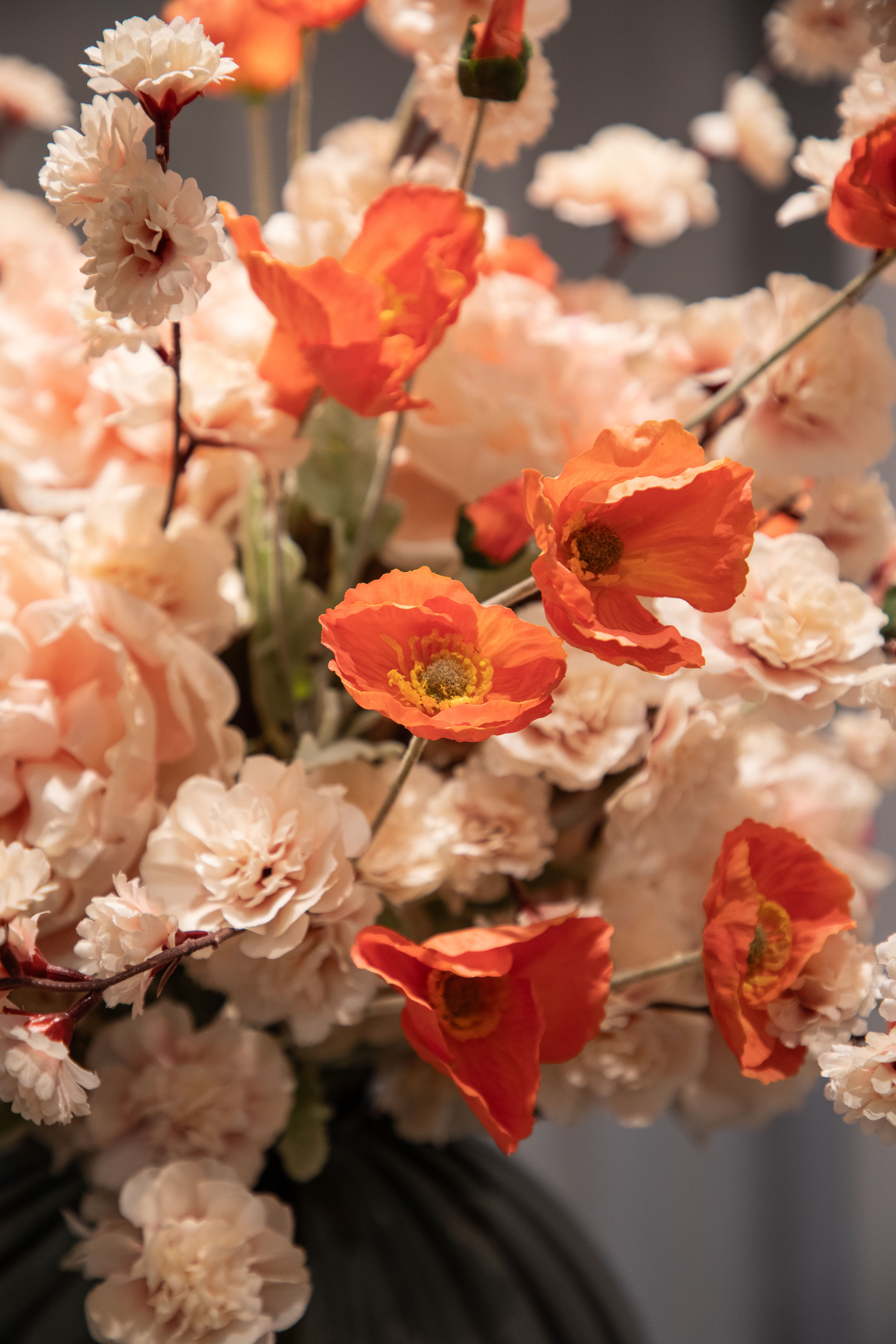 Nærbillede af orange valmuer, der er del af en større kunstig buket i sarte rose og orange nuancer, der er placeret i en grå glas vase.