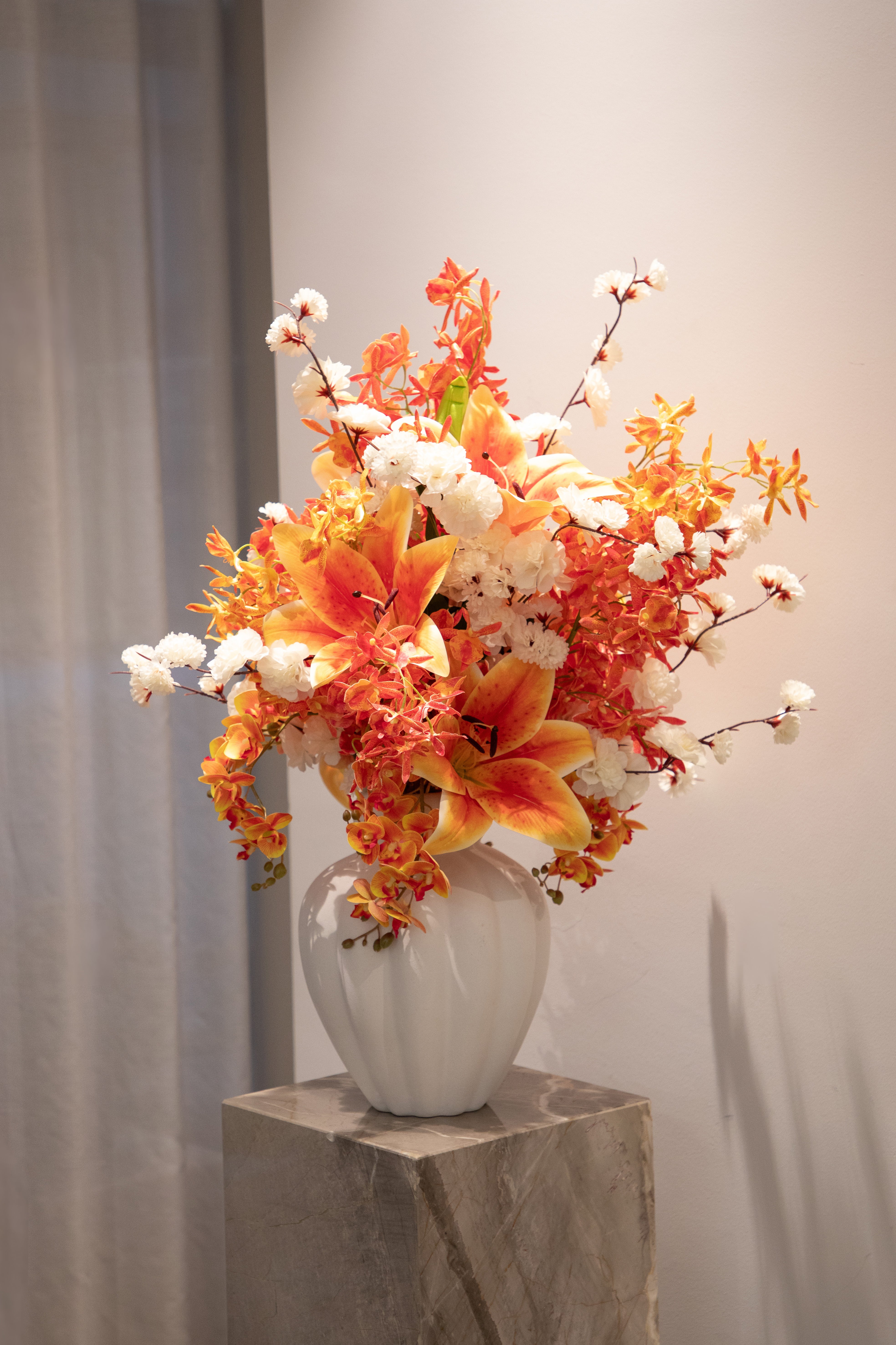 Mellem kunstig buket i hvide og orange farver, der står i en hvid keramik vase ovenpå en marmor piedestal. Baggrunden er hvid.