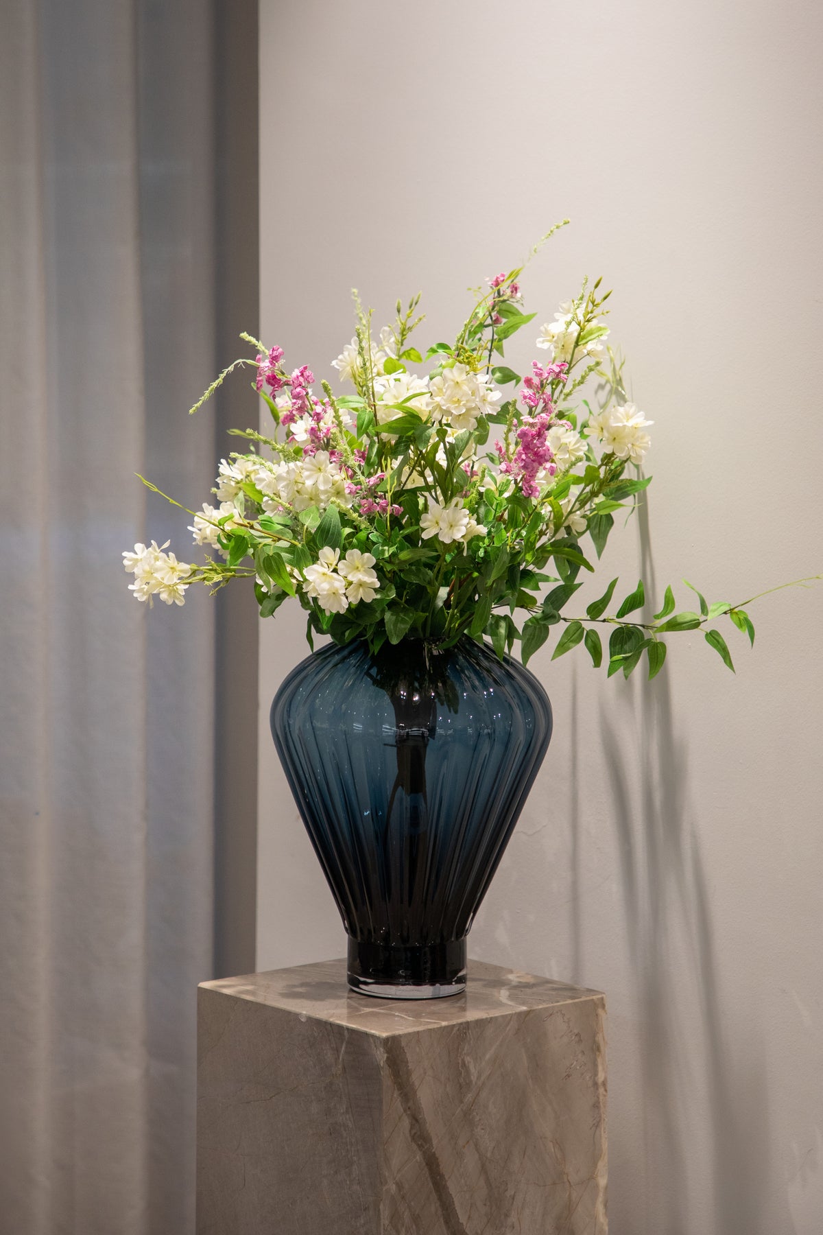 Stort sommerlig kunstig buket i grønne, hvide og rosa nuancer, der står i en mørkere blå glas vase ovenpå en marmor piedestal.