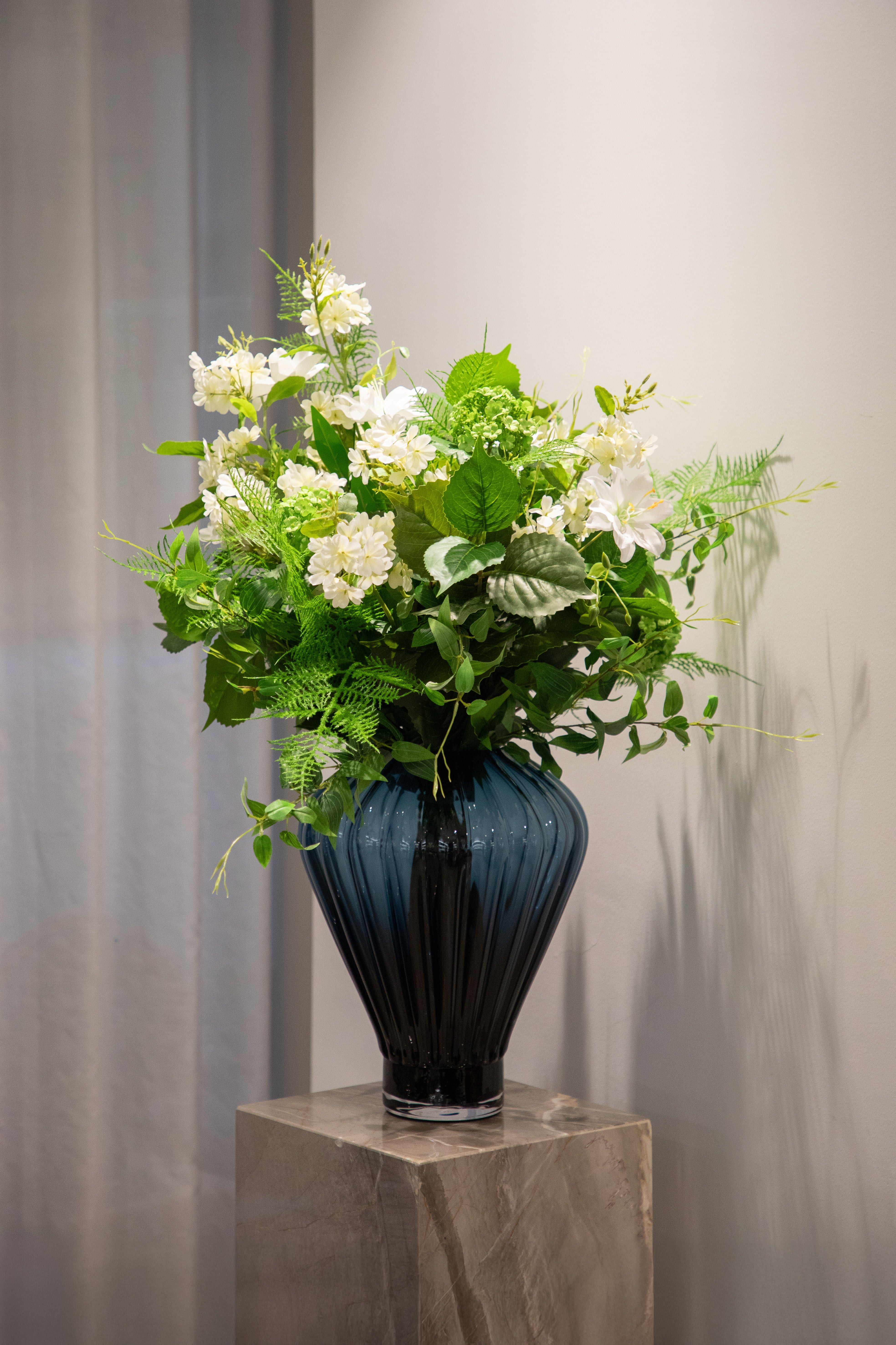 Stor frisk kunstig buket i grønne og hvide nuancer, der står i en stor blå glas vase ovenpå en marmor piedestal.