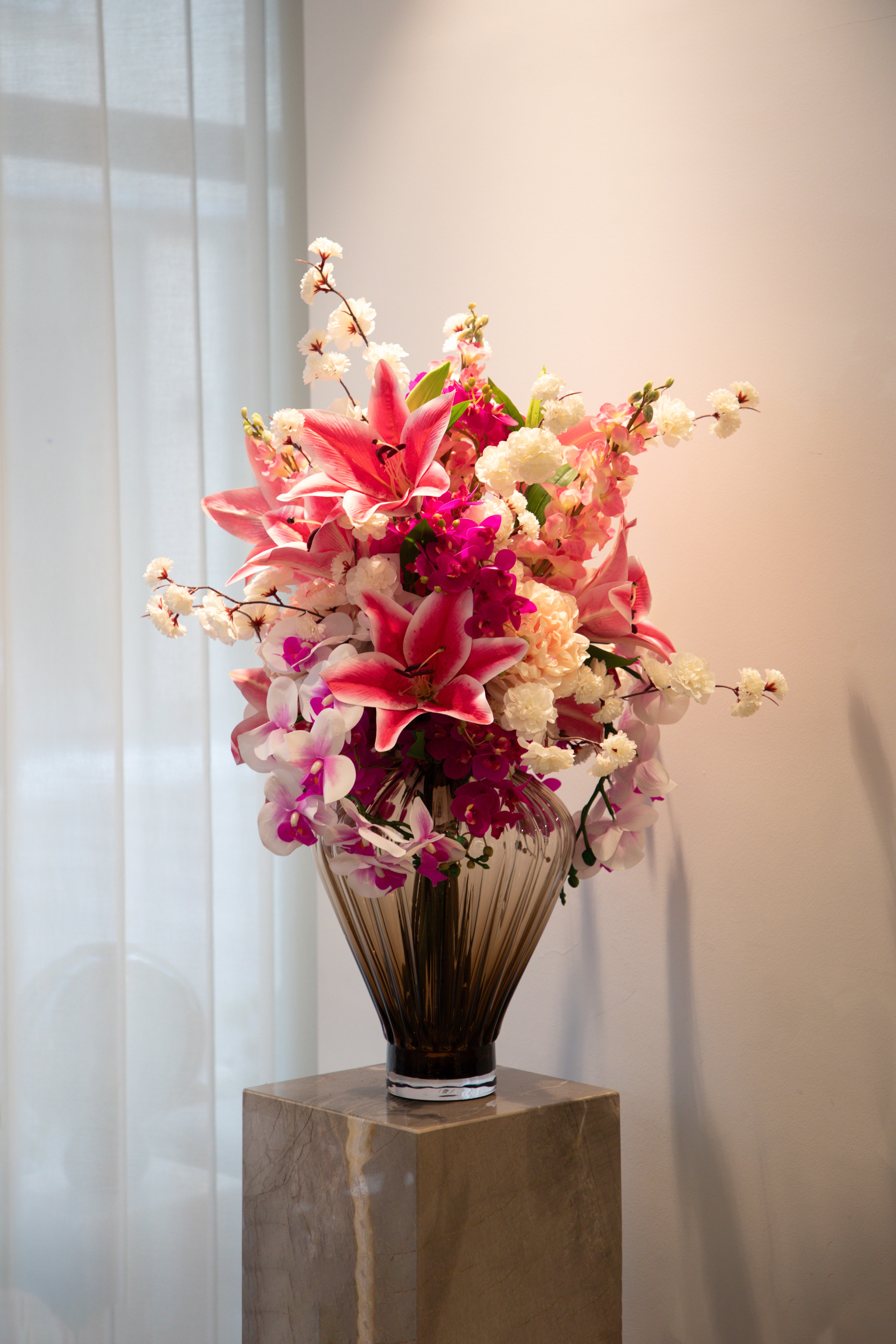 Stor romantisk kunstig buket i lyserøde, rosa og hvide nuancer, der står i en brun/røgfarvede glas vase ovenpå en marmor piedestal.