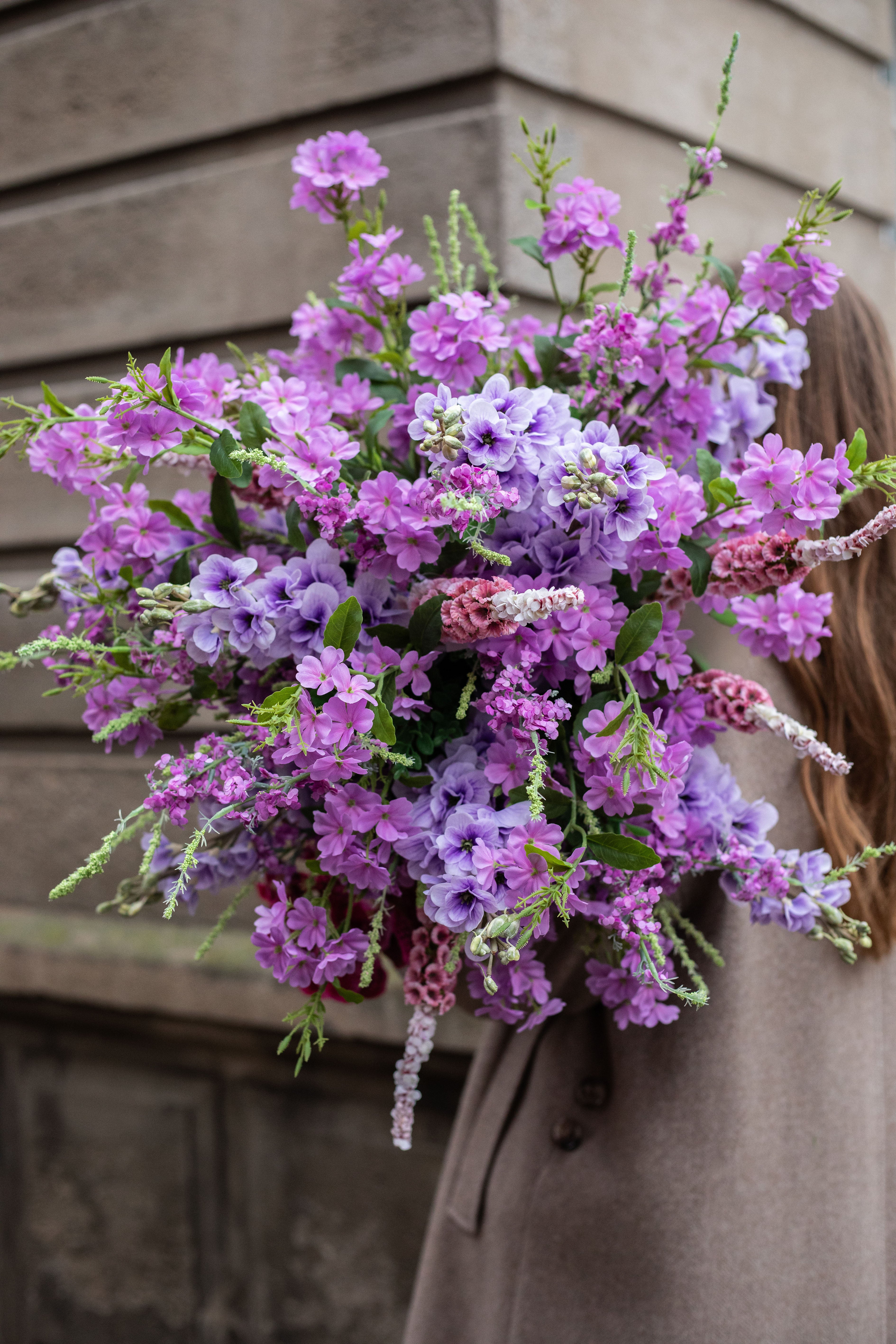 Nærbillede af stor kunstig buket i lilla og rosa nuancer. Buketten holdes af en pige med brunt hår og beige frakke. Billedet er taget udenfor, foran en beige mur.