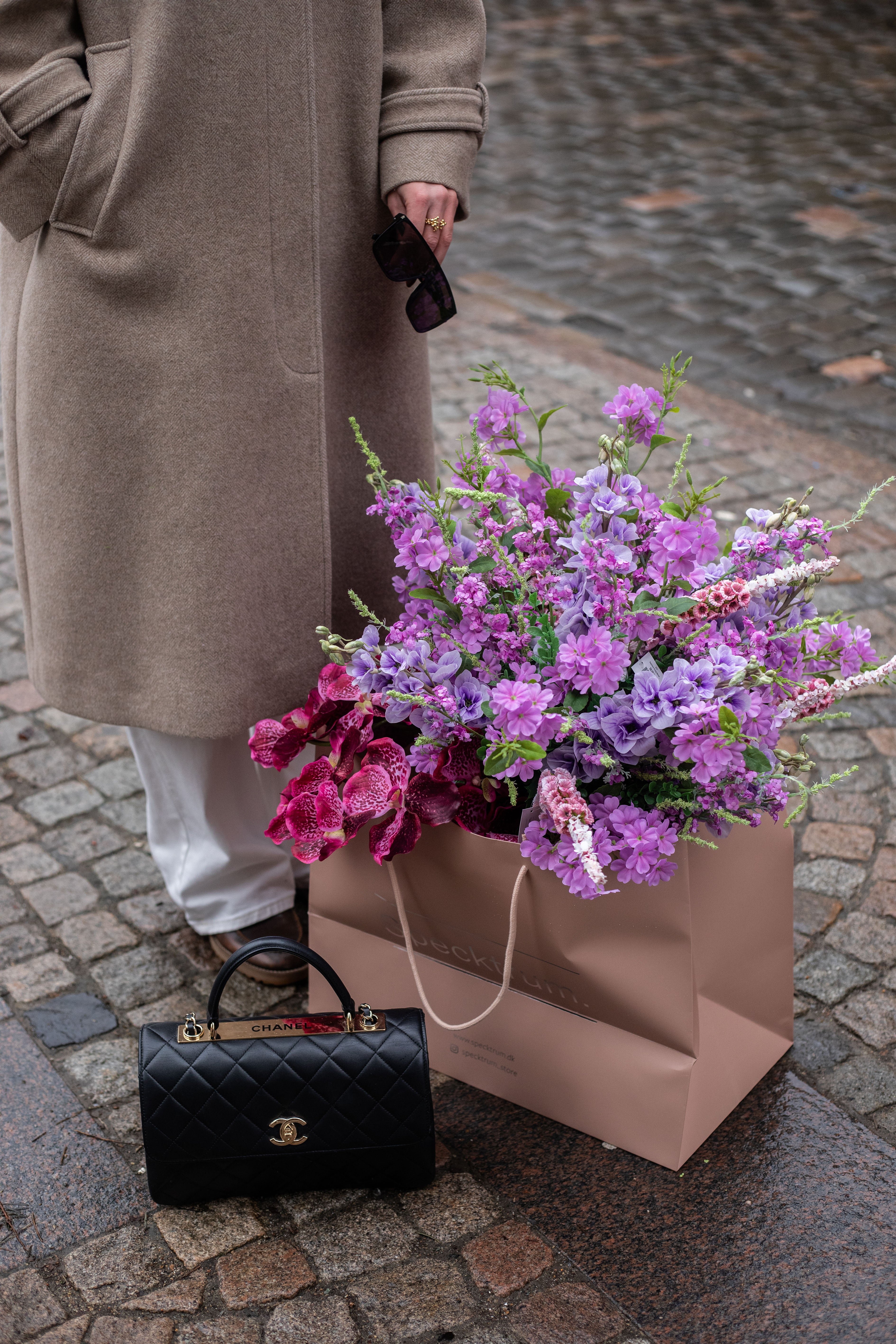 Stor kunstig buket i lilla og rose nuancer, der står i en lyserød Specktrum pose på jorden. Bagved står en pige med hvide jeans og beige frakke. Foran posen står en sort Chanel taske.