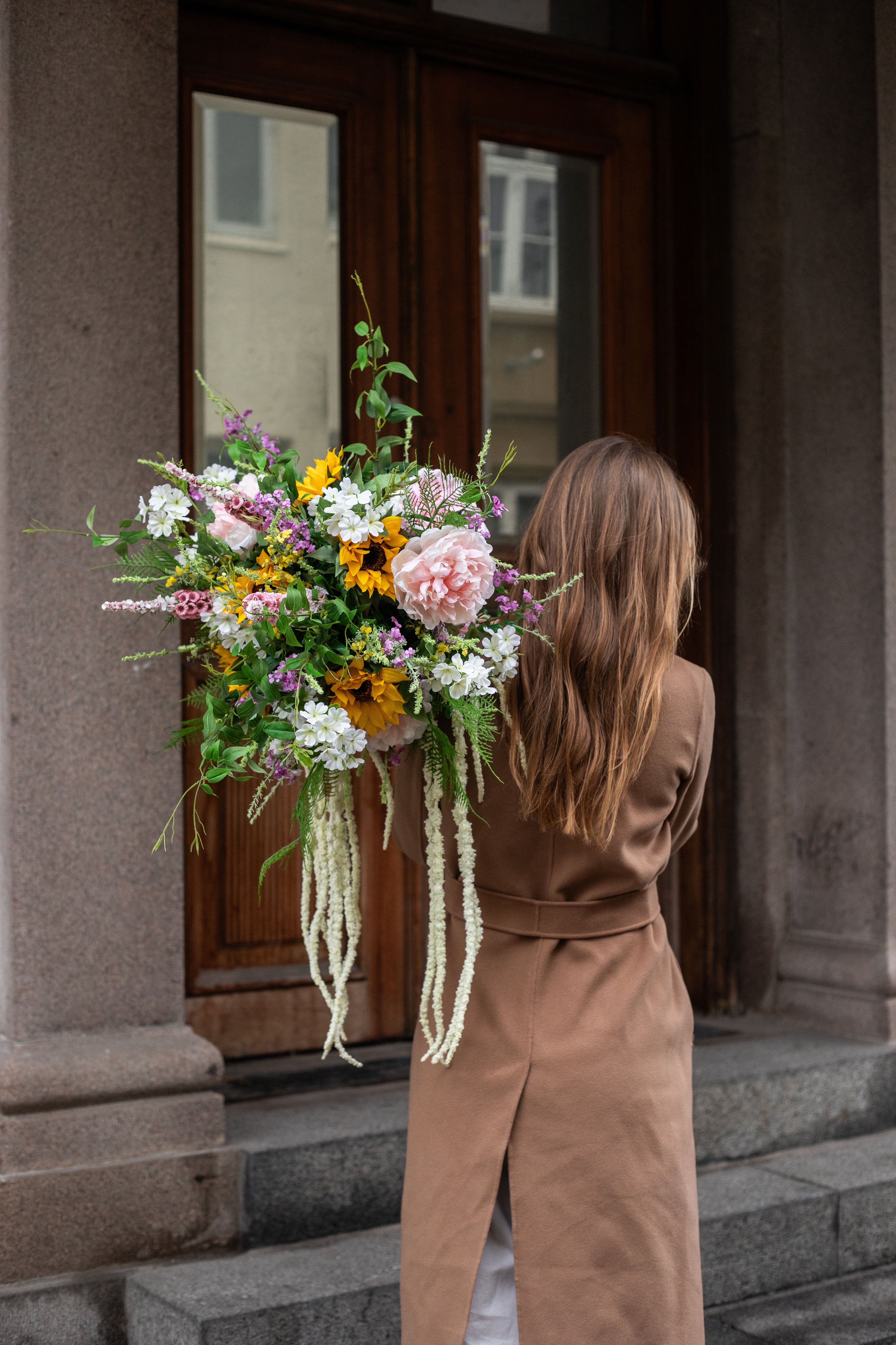 Pige der står med ryggen til. Hun har langt brunt hår og en brun frakke på. Over skulderen holder hun en stor kunstig buket fra Specktrum i gule, rose, grønne og hvide nuancer. Billedet er taget foran en grå bygning med brun dør.