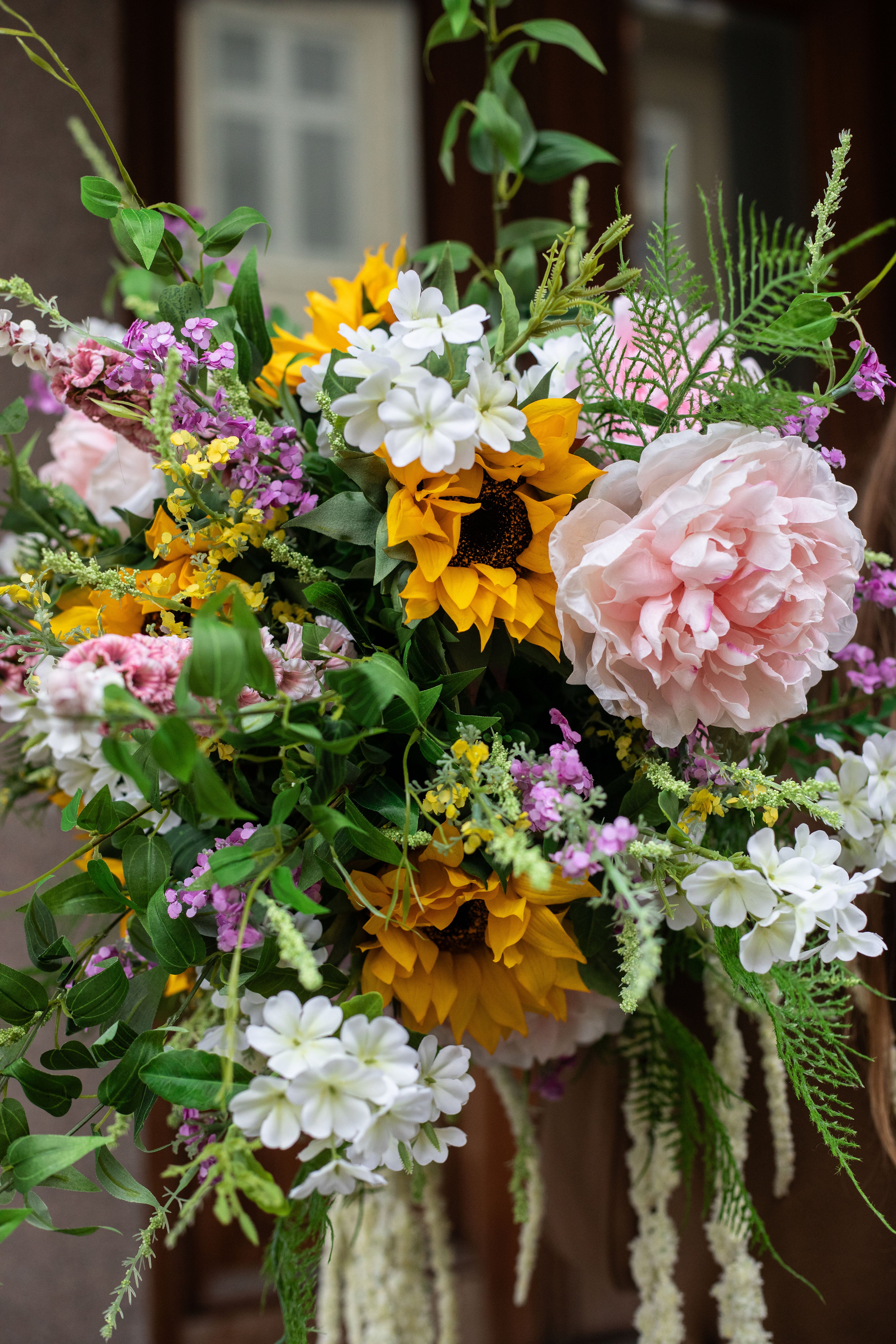 Nærbillede af en stor kunstig buket i grønne, gule, rose og hvide nuancer. Buketten holdes af en pige med brun frakke, op foran hendes hoved.