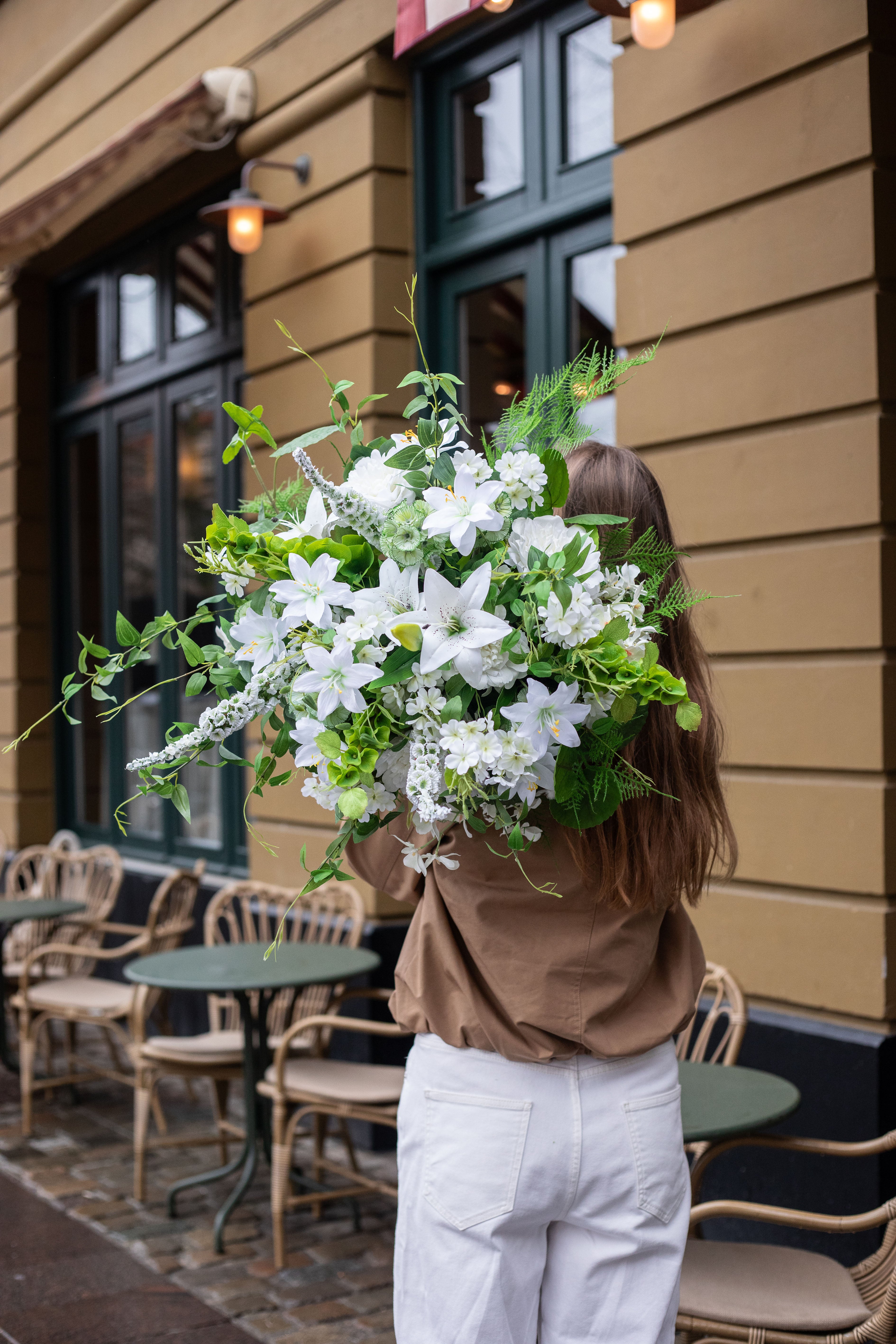 Pige der står med ryggen til kameraet. Hun har langt brunt hår, beige jakke og hvide jeans på. Over skulderen holder hun en stor kunstig buket i hvide og grønne nuancer. Baggrunden er en orange bygning, foran står cafeborde og stole.