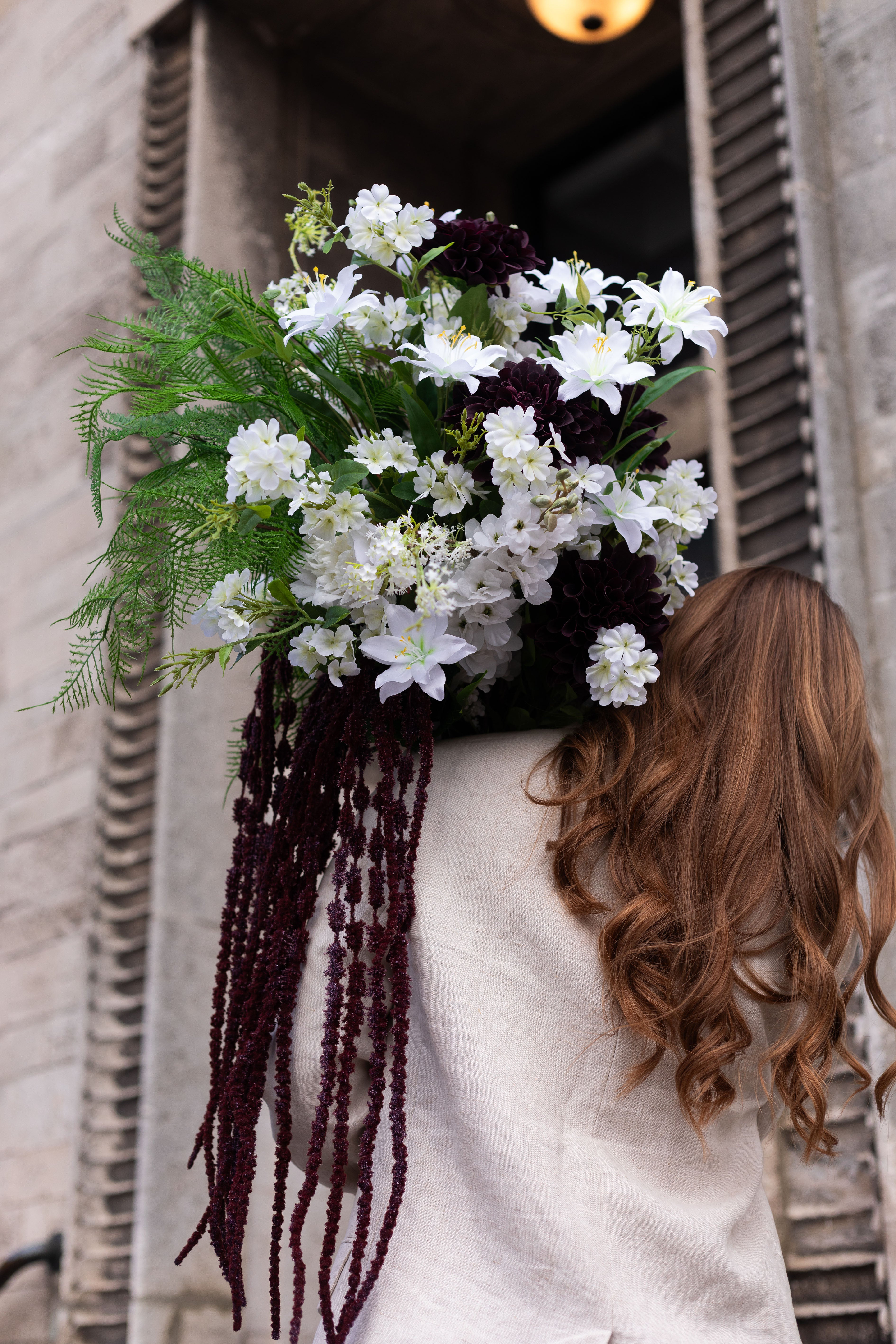 Pige med ryggen til, hun har langt brunt hår og beige blazer på. Over skuldren holder hun en stor kunstig buket i hvide, bordeaux og grønne nuancer fra Specktrum. Baggrunden er en murstens bygning i beige nuancer.