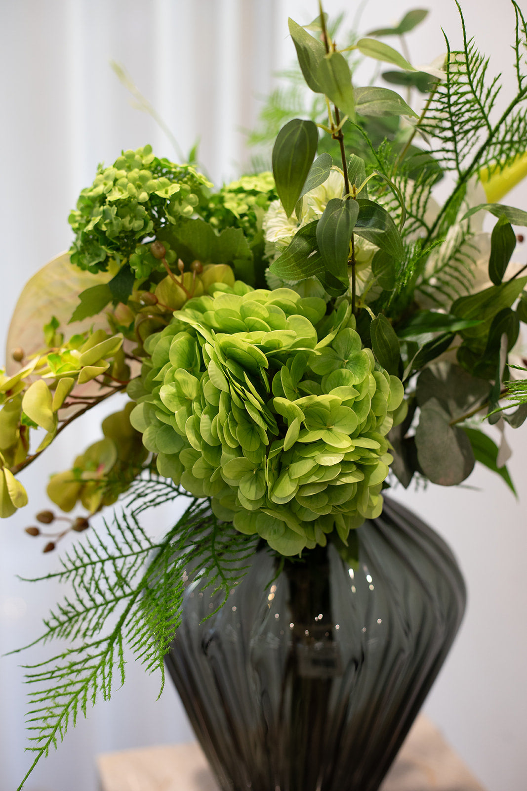 Nærbillede af kunstig buket i grønne nuancer, stor hortensia i fokus. Buketten er placeret i en grå glas vase.