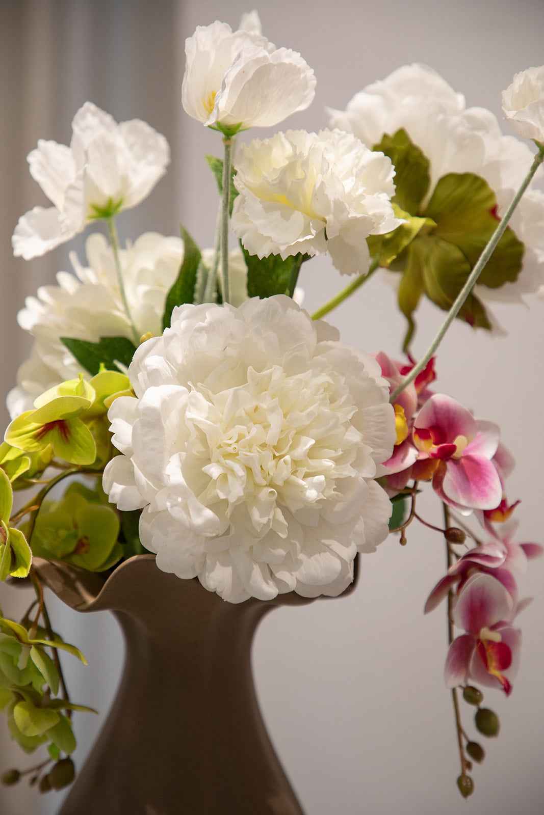 Nærbillede af en buket af kunstige blomster i hvide, grønne og rosa nuancer. Placeret i en brun keramikvase.