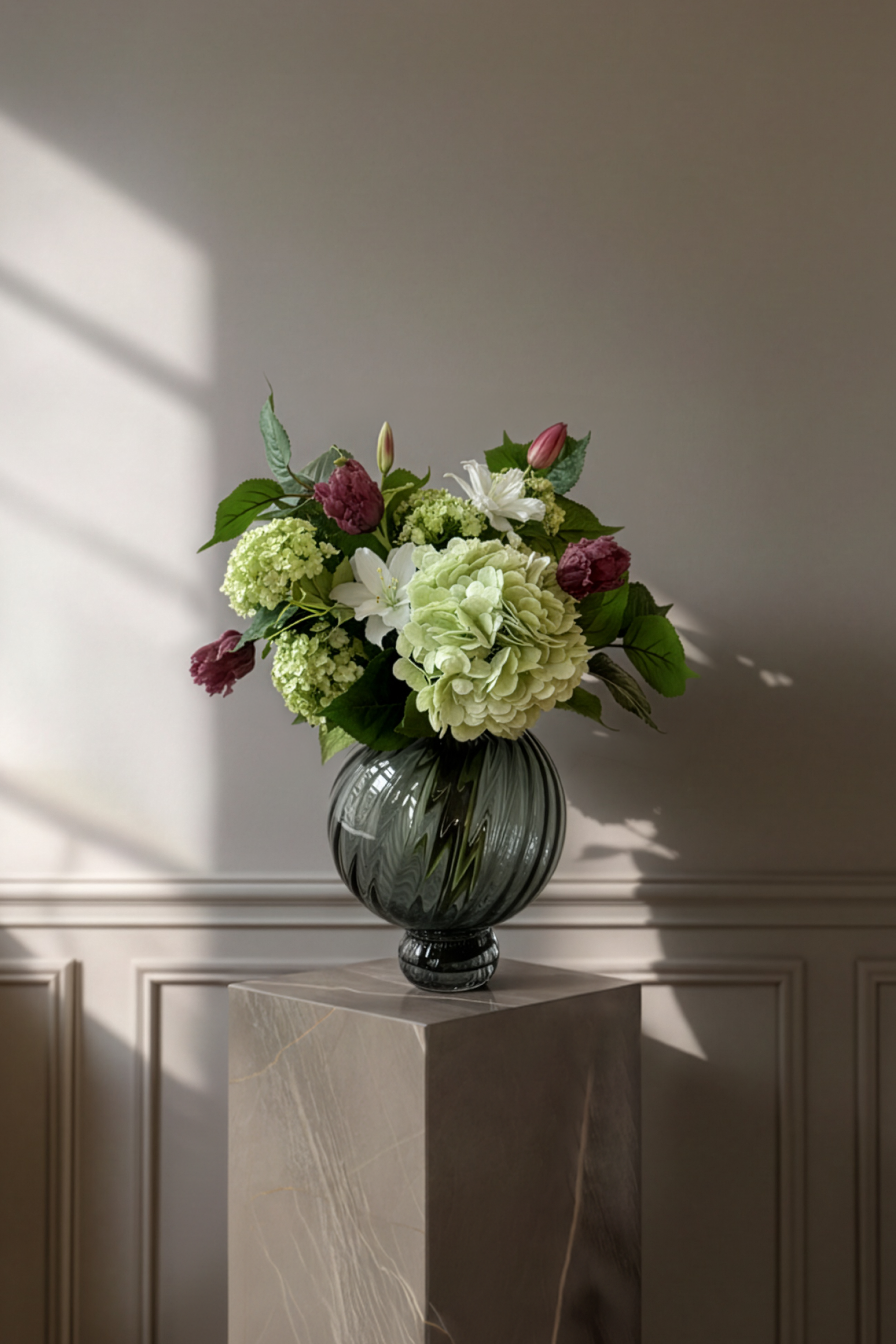 Basisbuket i grønne nuancer, piftet op med smukke tulipaner i mørkelilla. Buketten er stylet i en rund glasvase på en marmor pedestal.