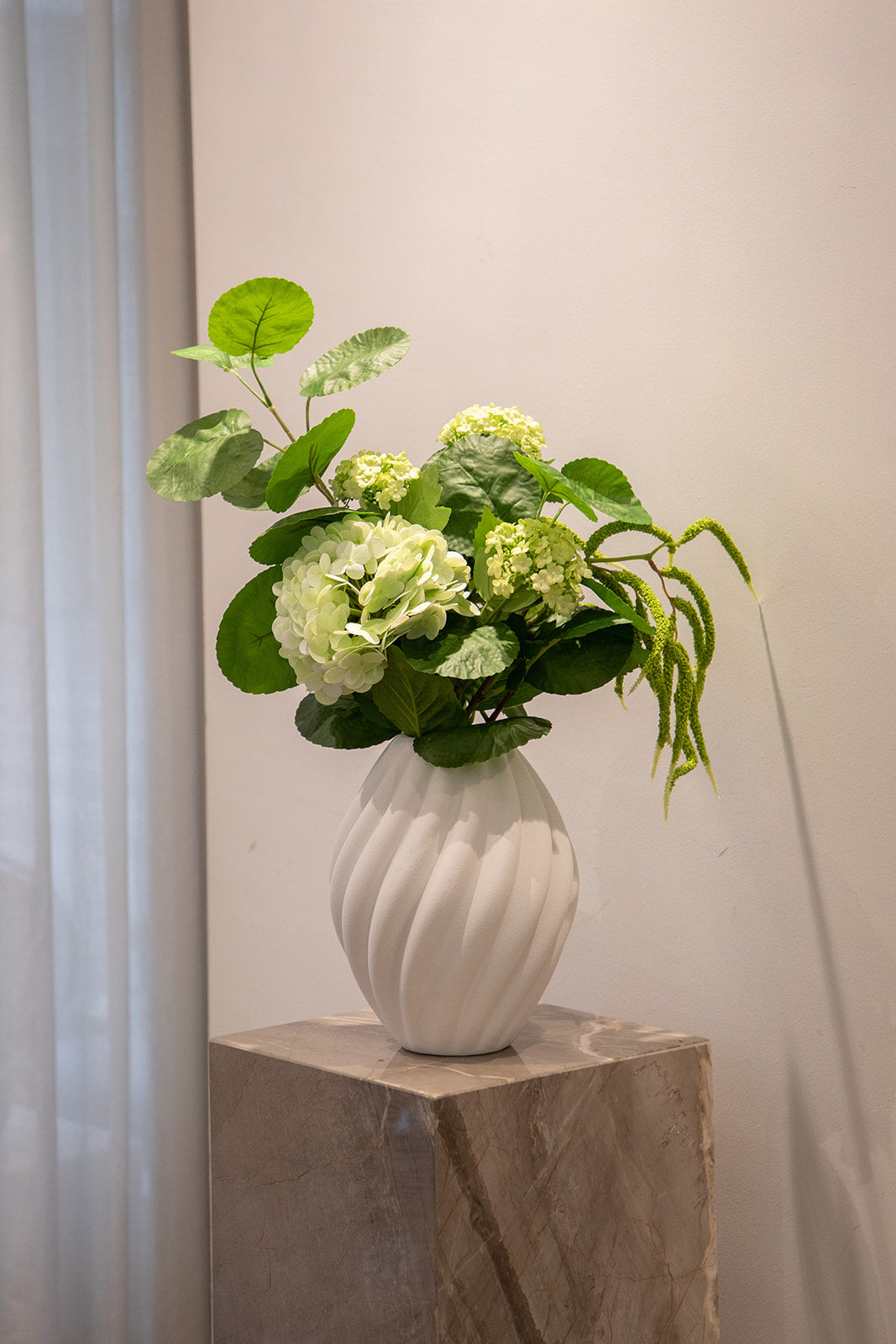 Basis kunstig buket i grønne nuancer, placeret i en hvid keramik vase. der står på en marmor piedestal.