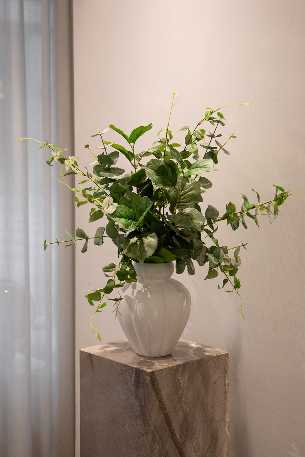 Foto af en kunstig blomster buket i grønne nuancer, der står i en hvid keramik vase. Placeret oven på en marmor piedestal.