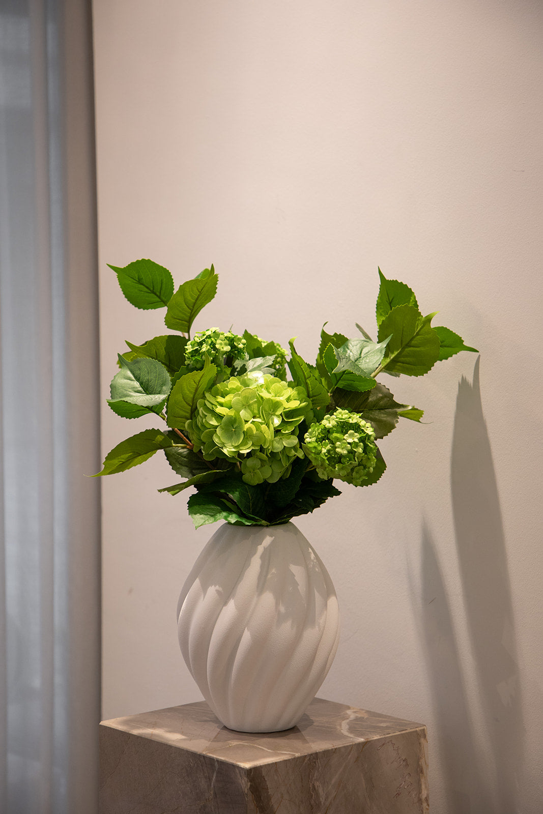 Kunstig buket i grønne nuancer, der er placeret i en hvid keramik vase, og står på en marmor piedestal.