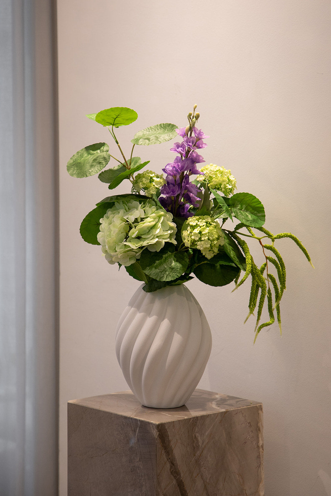 En kunstig basis blomster buket i grønne nuancer, der er tilføjet en lilla stilk. Buketten er placeret i en hvid keramik vase, der står på en marmor piedestal.