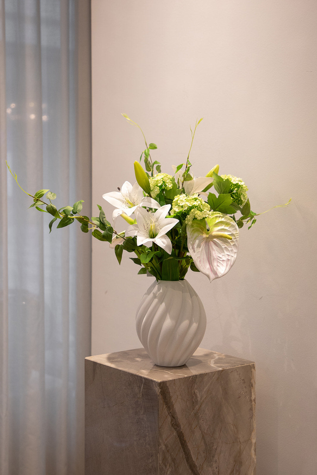Foto af kunstige blomster i grønne og hvide nuancer, placeret i en hvid keramik vase, der står på en marmor piedestal.