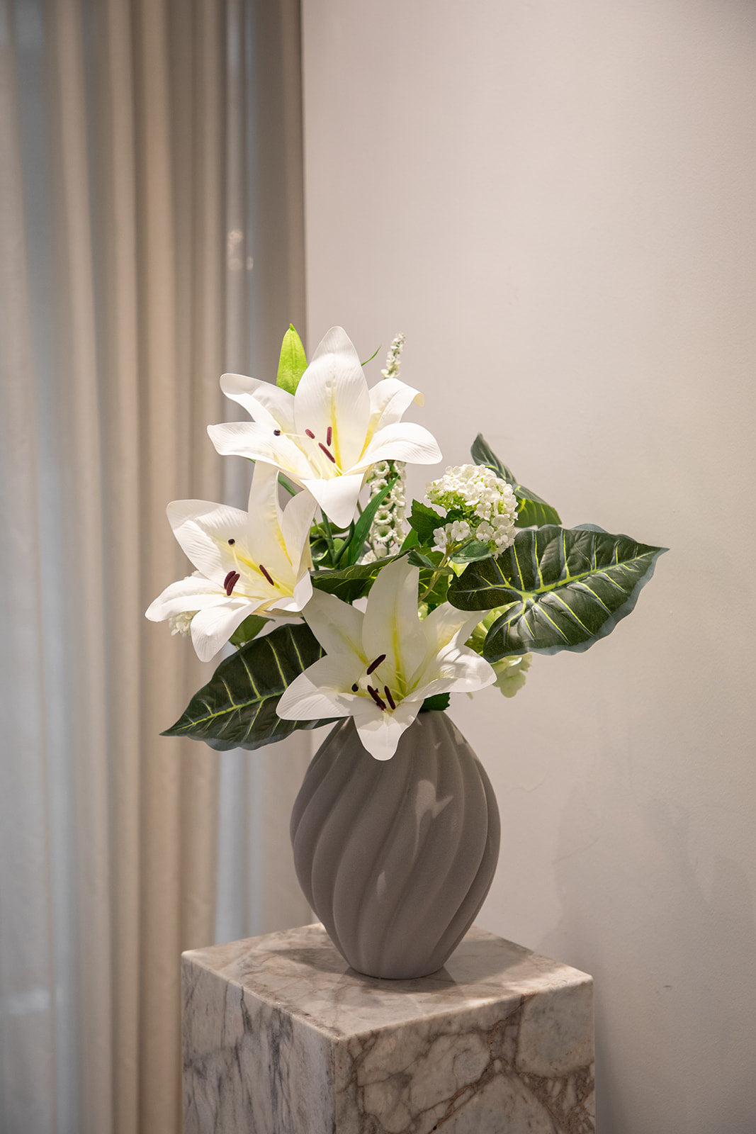 Buket af kunstige blomster i hvide og grønne nuancer. Placeret i en keramikvase, der står oven på en marmorpedestal.