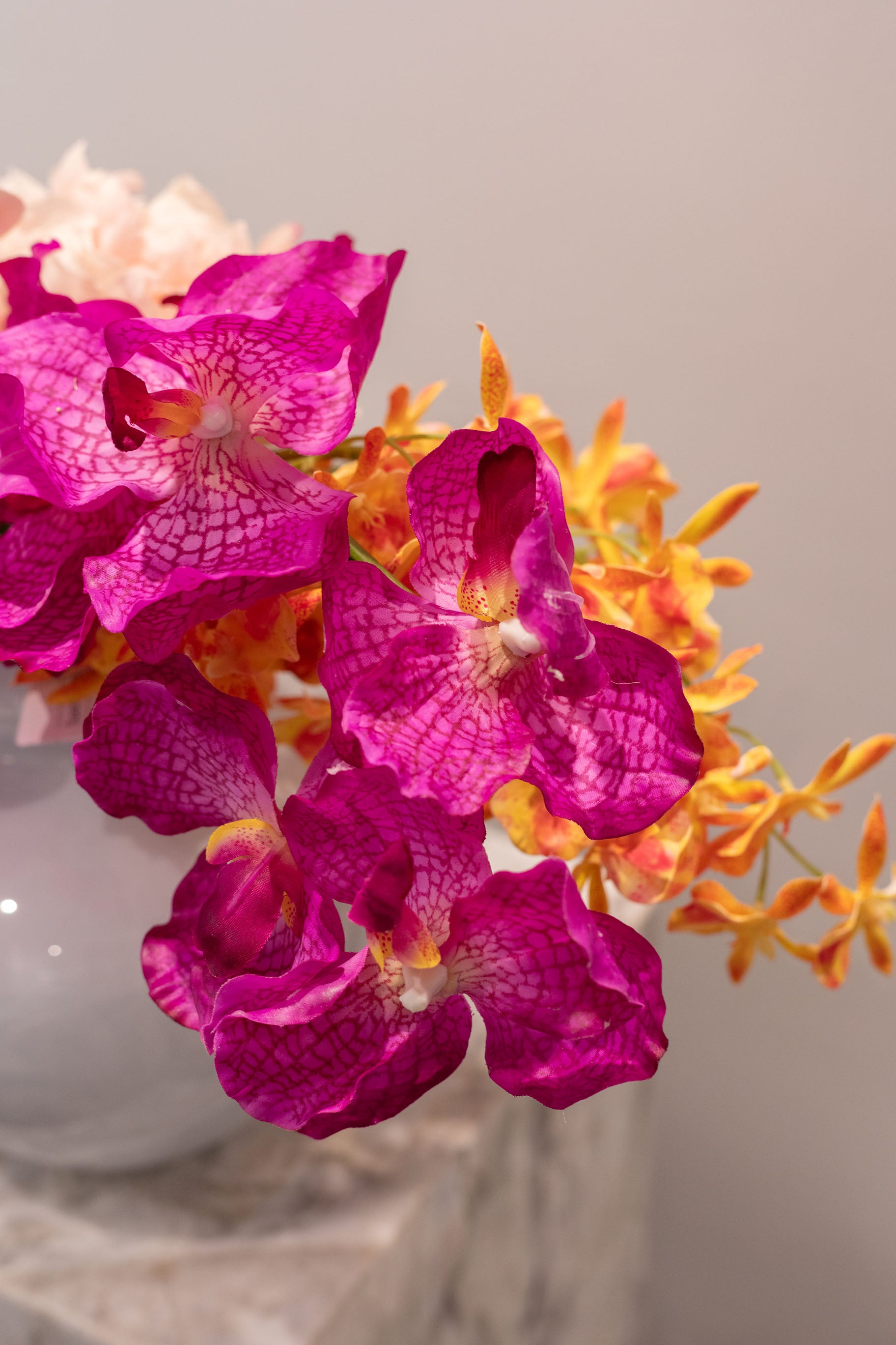 Nærbillede af flerhovedet orkidé i lilla/lyserød. En del af en buket i orange, grøn og rosa nuancer. Placeret i en blå glasvase, der står på en marmor pedestal.