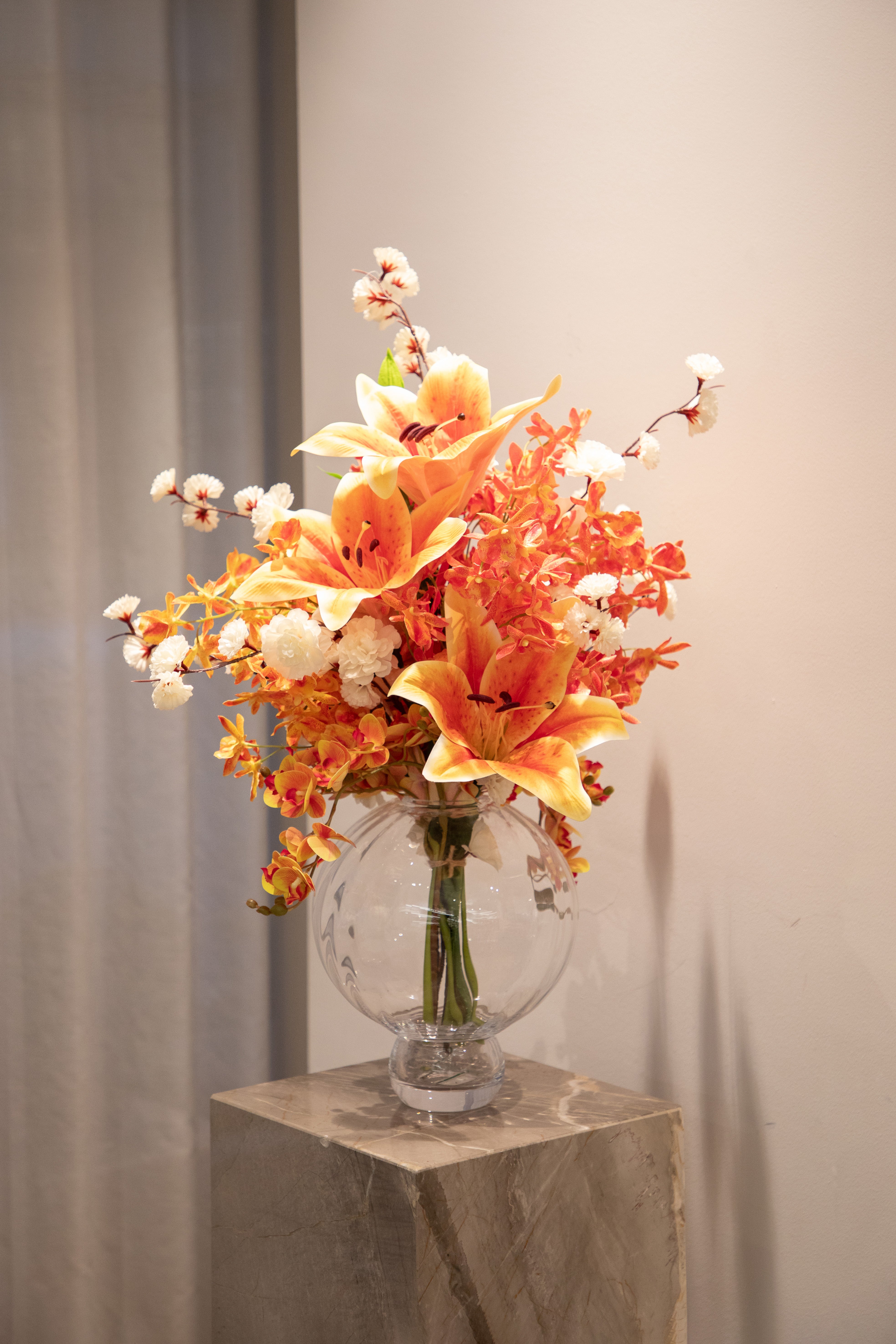 Kunstig buket i orange og hvide farver, der står i en gennemsigtig rund glas vase ovenpå en marmor piedestal.