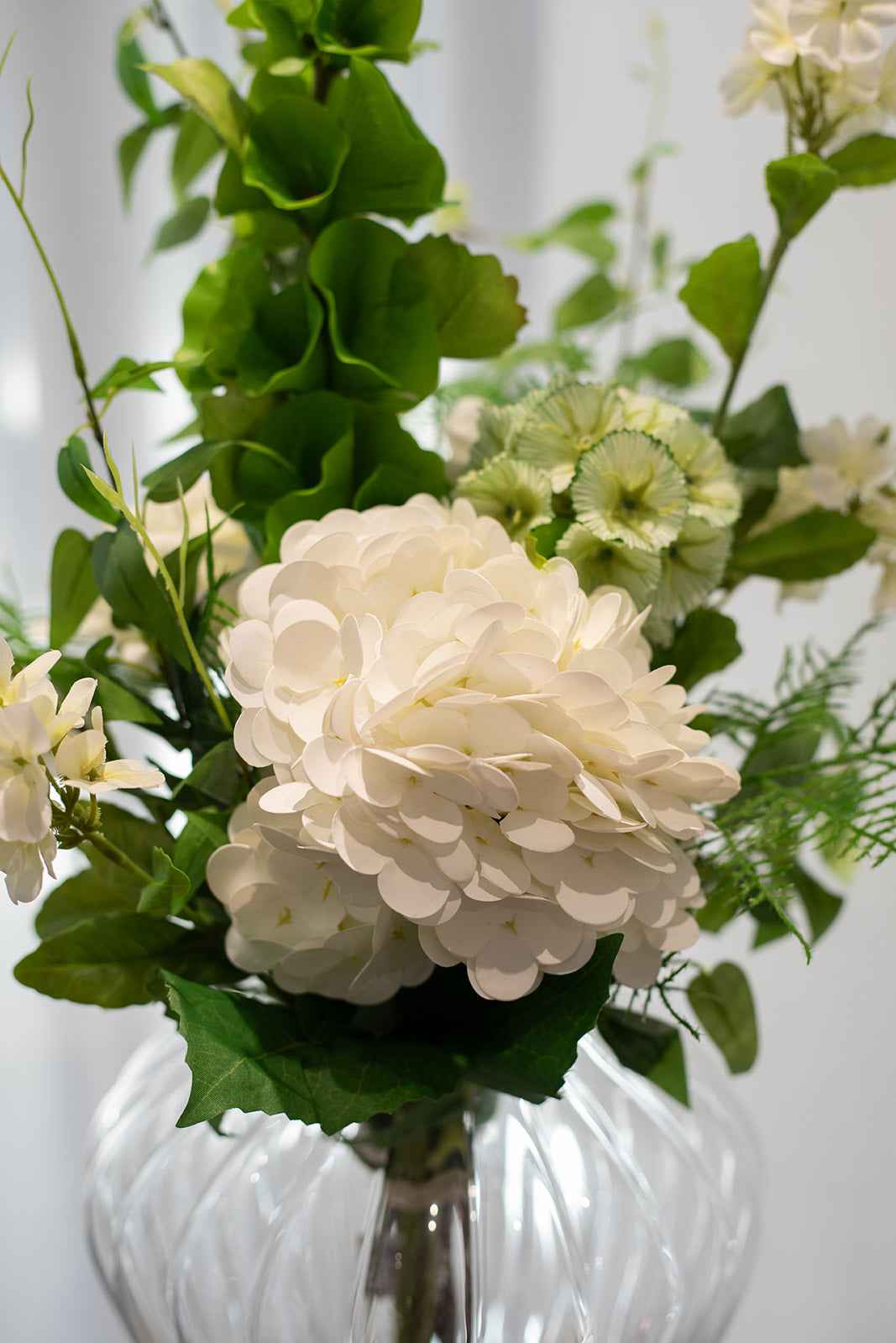 Nærbillede af kunstig buket i grønne og hvide nuancer, placeret i en gennemsigtig glas vase.