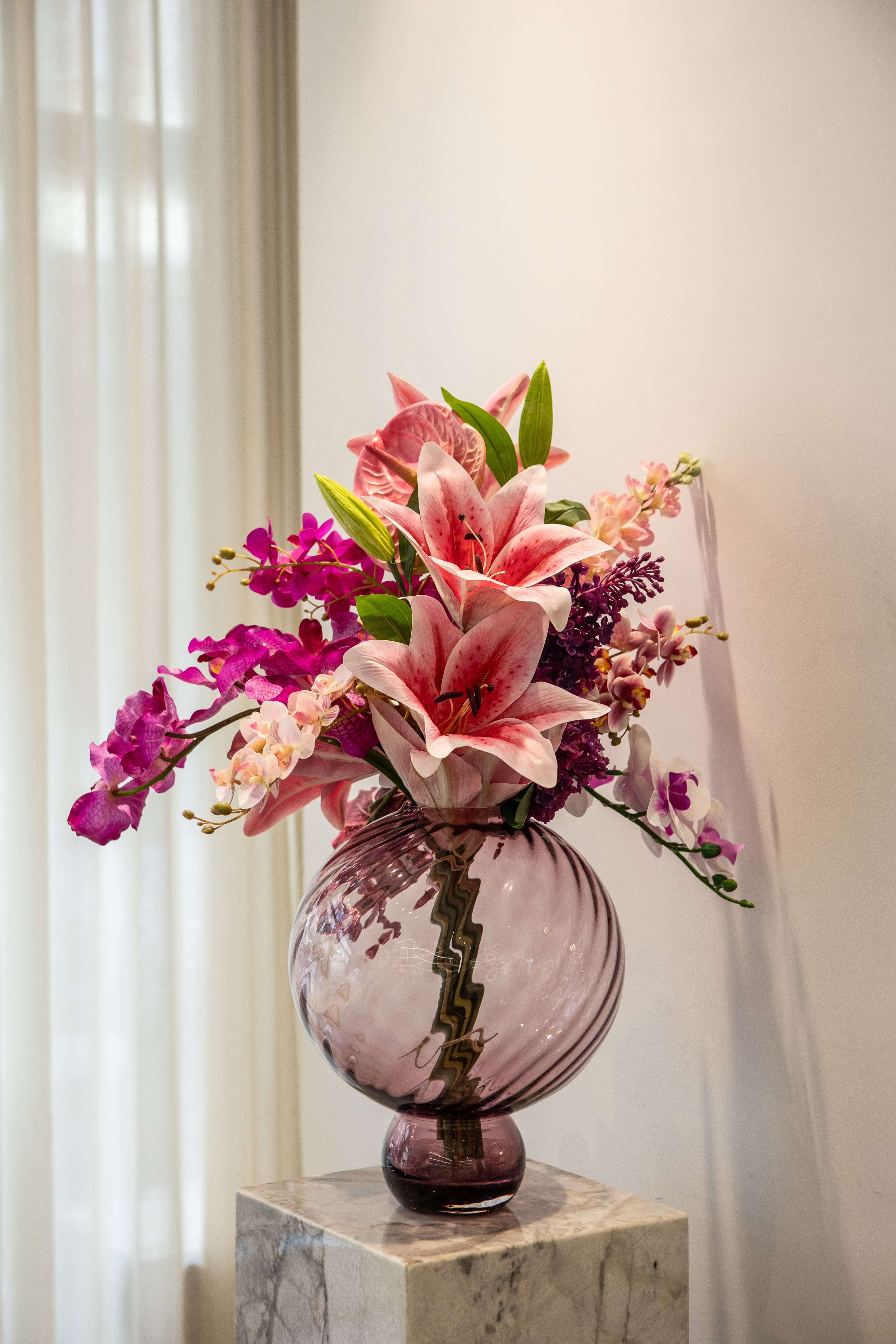 Stor buket af kunstige blomster i lilla og lyserøde nuancer i en stor plum farvet glasvase, der er placeret på en marmorpedestal.