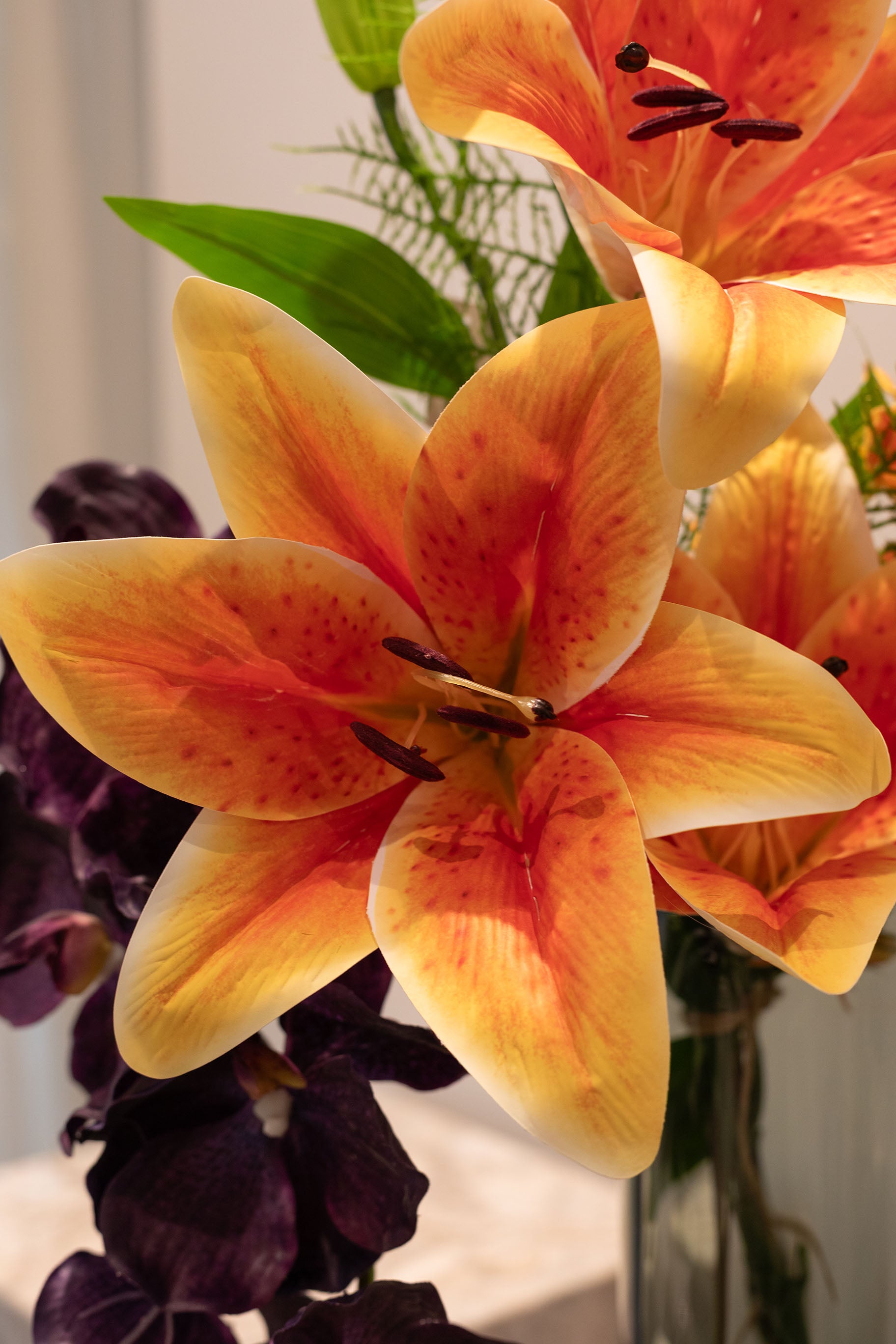 Nærbillede af en stor orange lilje, en del af en buket af kunstige blomster i mørke lilla og grønne nuancer.