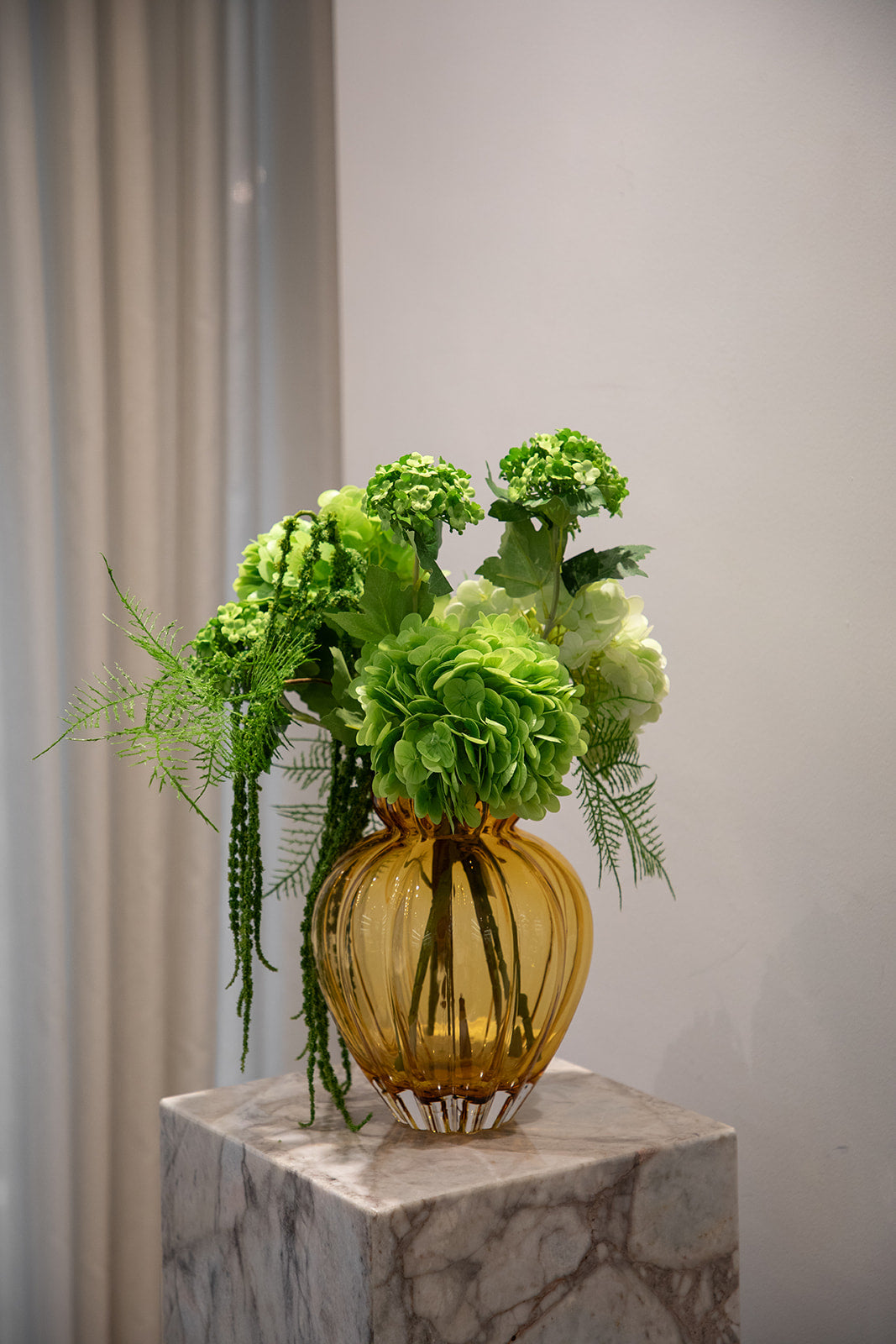 Billede af buket af kunstige blomster i grønne nuancer. Placeret i en orange farvede glasvase, der står på en marmor pedestal.