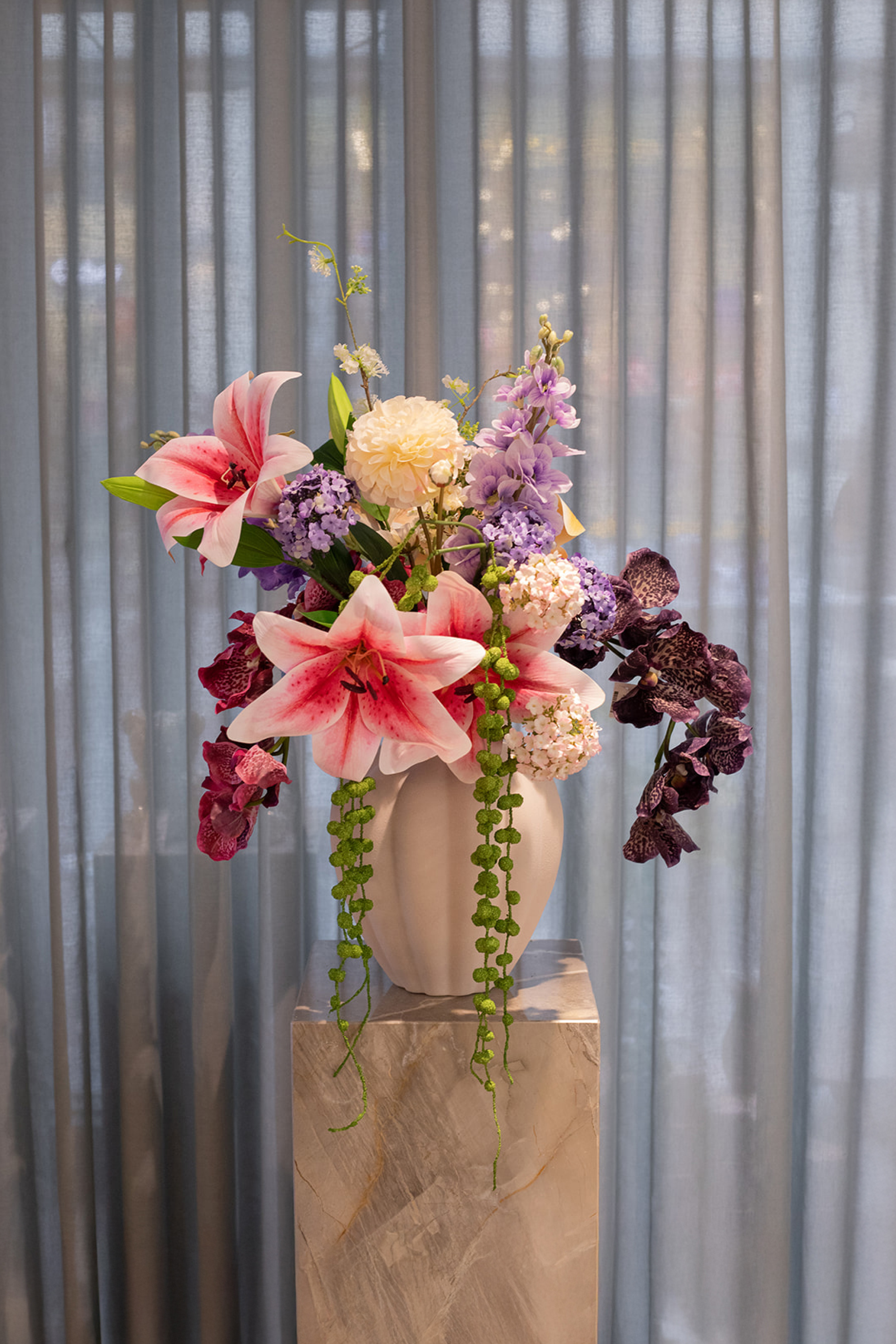 En lys vase med en farverig buket af lilla, orange, pink og mørkelilla blomster samt grønne detaljer, placeret på en marmorpiedestal foran lyse gardiner.