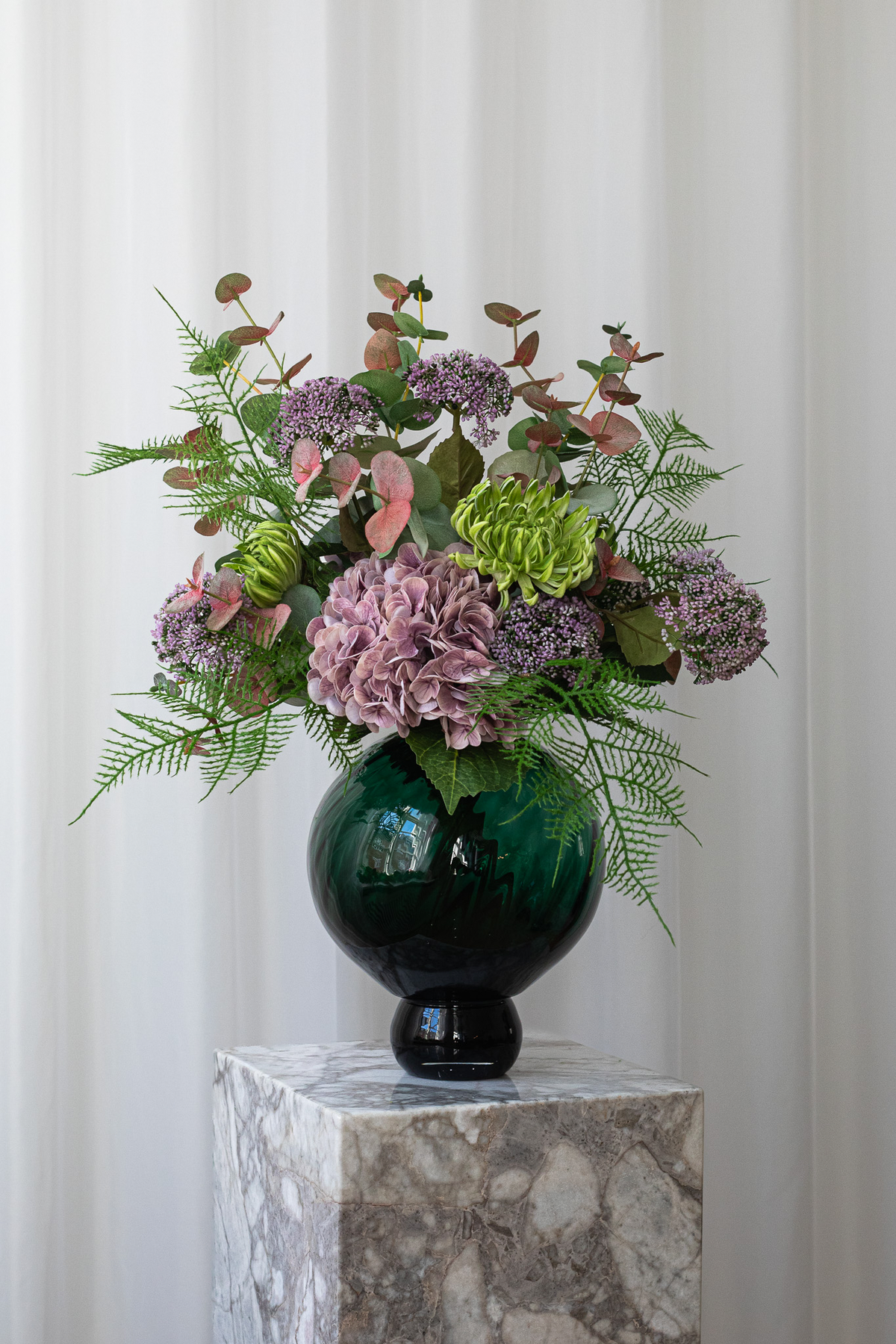 En smuk, kunstig buket med lyselilla hortensia, lilla bærgrene, bregner, eukalyptusgrene og andet grønt. Buketten står skulpturelt og naturtro i en grøn glasvase.