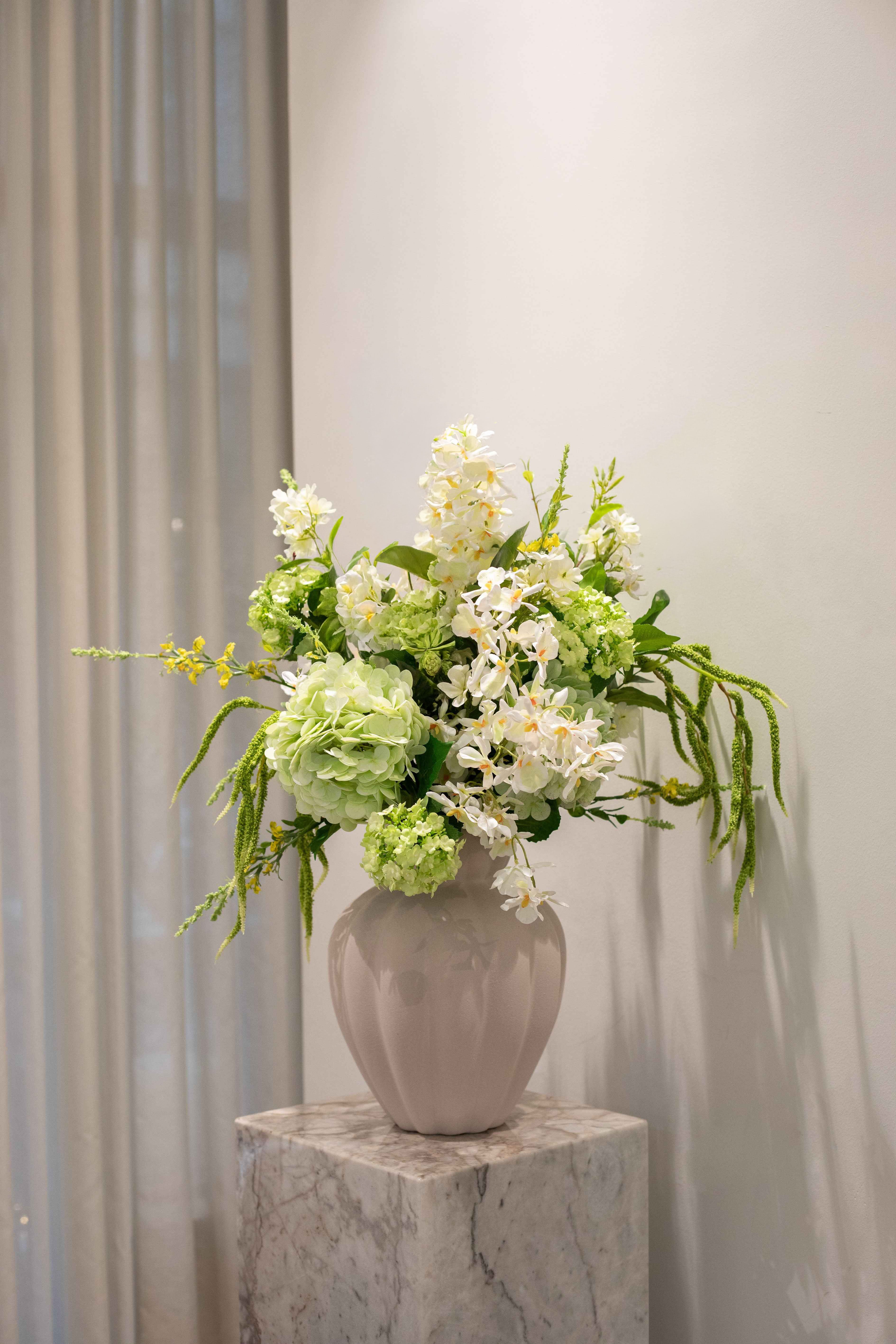 En stor og luftig buket i hvide, grønne og lyse gule nuancer. Buketten er stylet i Penelope vasen i sandfarvet.