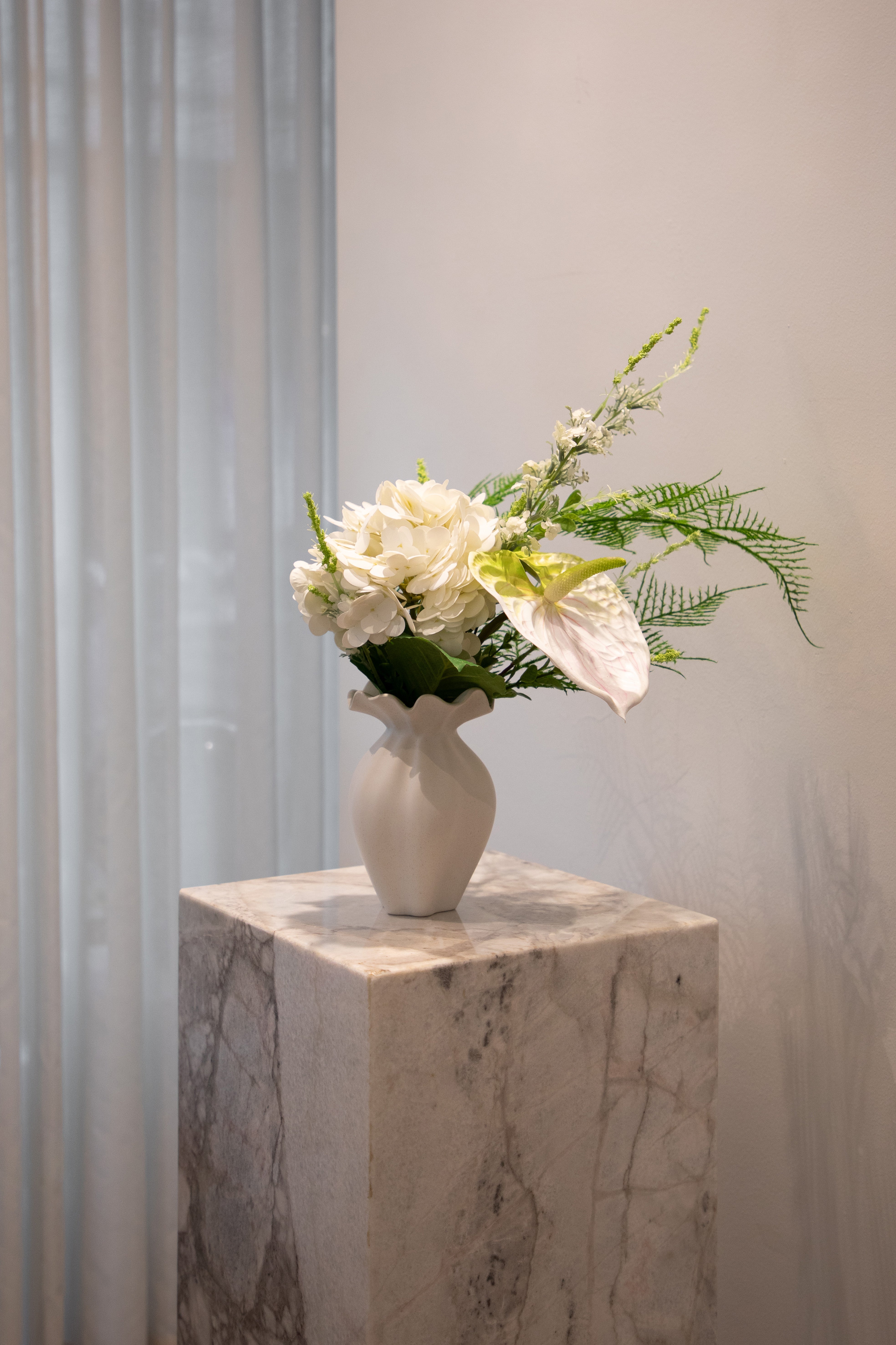 Kunstig buket i hvide og grønne nuancer, placeret i en hvid keramik vase, der står på en marmor pedestal.